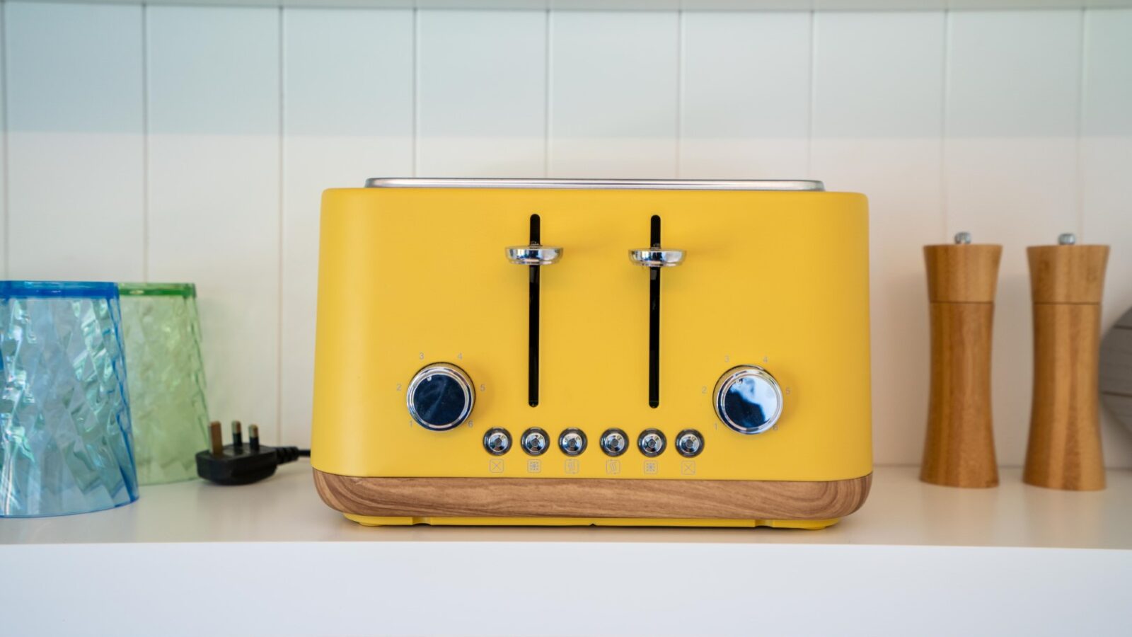 A yellow toaster with two slots, control buttons, and dials sits on a white countertop at Baxby Manor. To the left are two textured glass cups, and to the right are two wooden salt and pepper grinders. The wall behind is tiled in white.