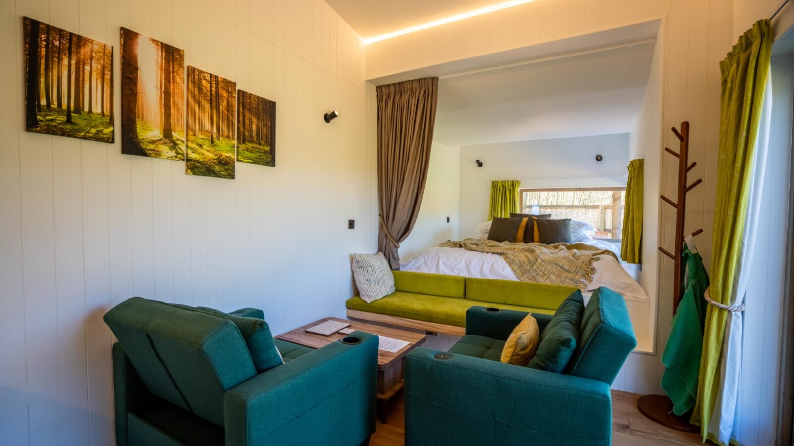 A cozy room at Baxby Manor featuring two green armchairs facing a wooden coffee table, with a bed in an alcove containing yellow and green bedding. A set of paintings depicting a forest decorates the white wall above the armchairs. Large windows provide natural light, enhancing the tranquil Kabinas feel.