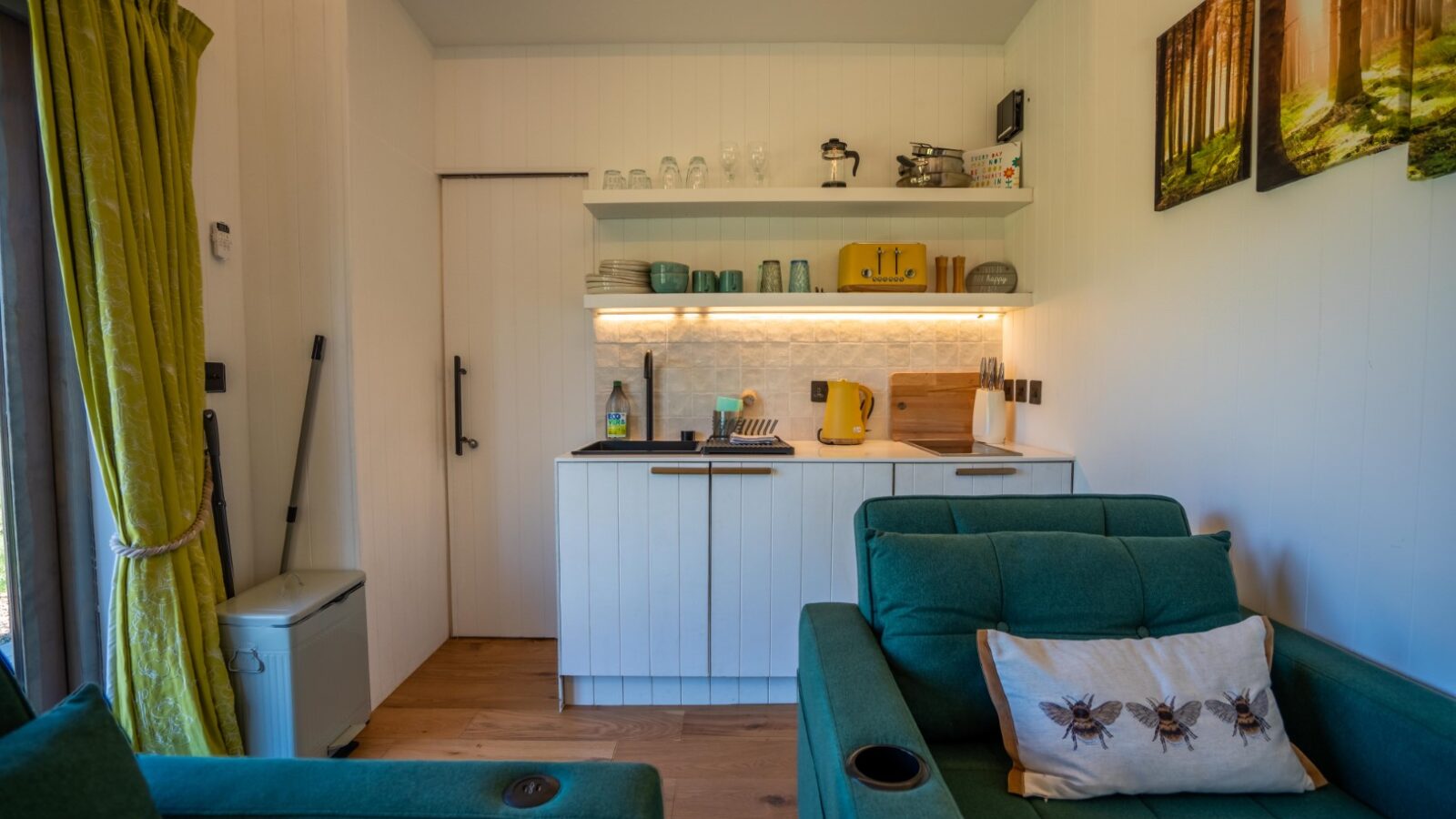 A cozy, small kitchen area with white cabinets and wooden countertops. There's a yellow toaster and matching kettle, shelves with glassware and dishes, a mini-fridge, and a green curtain by the window. A green armchair with a bee-themed pillow from Kabinas is in the foreground.