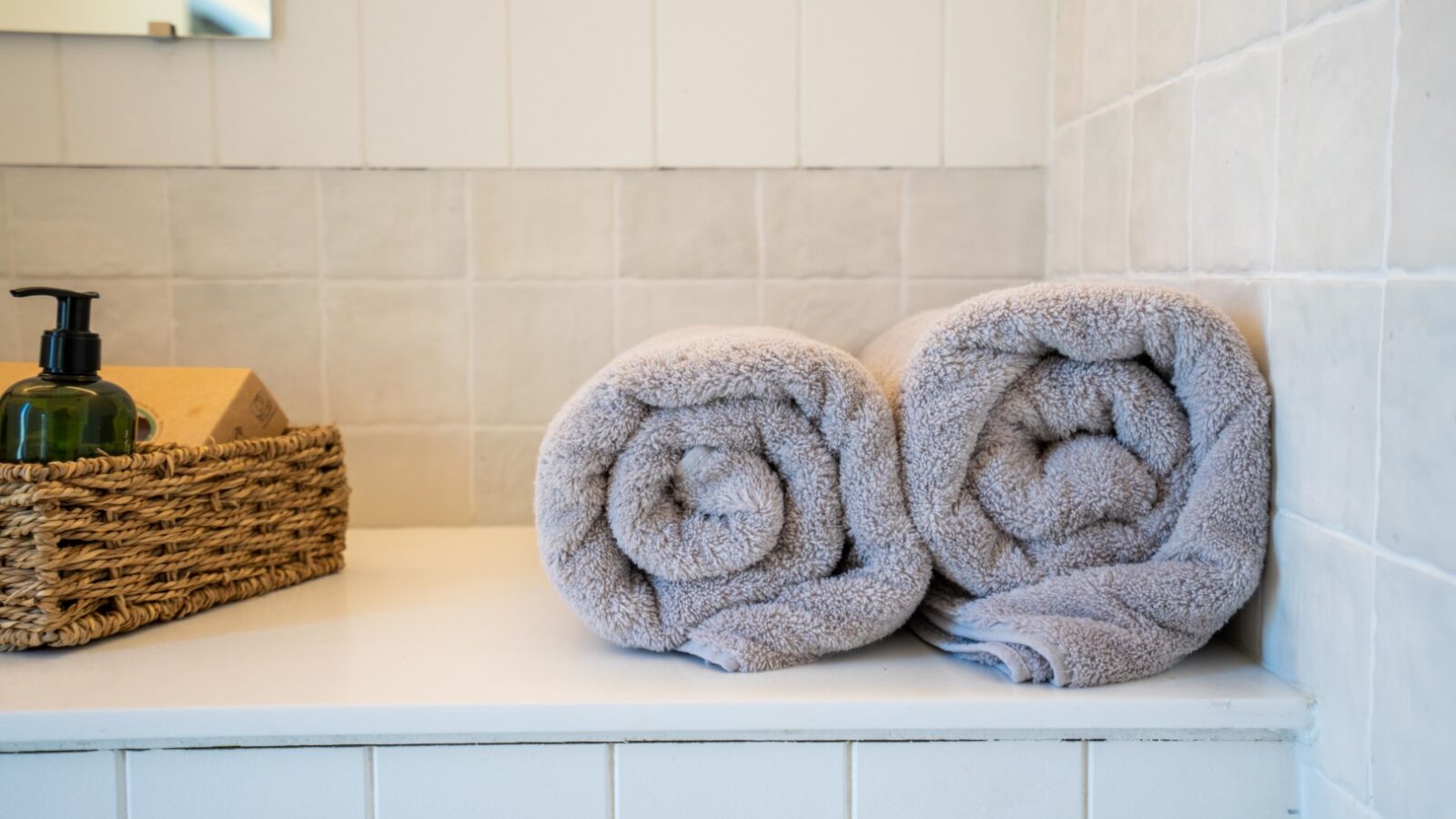 Two neatly rolled gray towels rest on a white bathroom countertop. A wicker basket containing a green liquid soap dispenser and a wooden box is situated to the left. The background, reminiscent of Baxby Manor, features white, textured, square tiles on the walls.