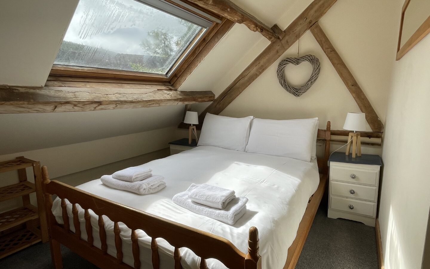 Nestled like the charming Trallwm Forest Cottages, this cozy attic bedroom features a skylight, wooden bed, pristine white bedding, and towels. A heart-shaped wicker decor graces the wall, adding to its homely allure.