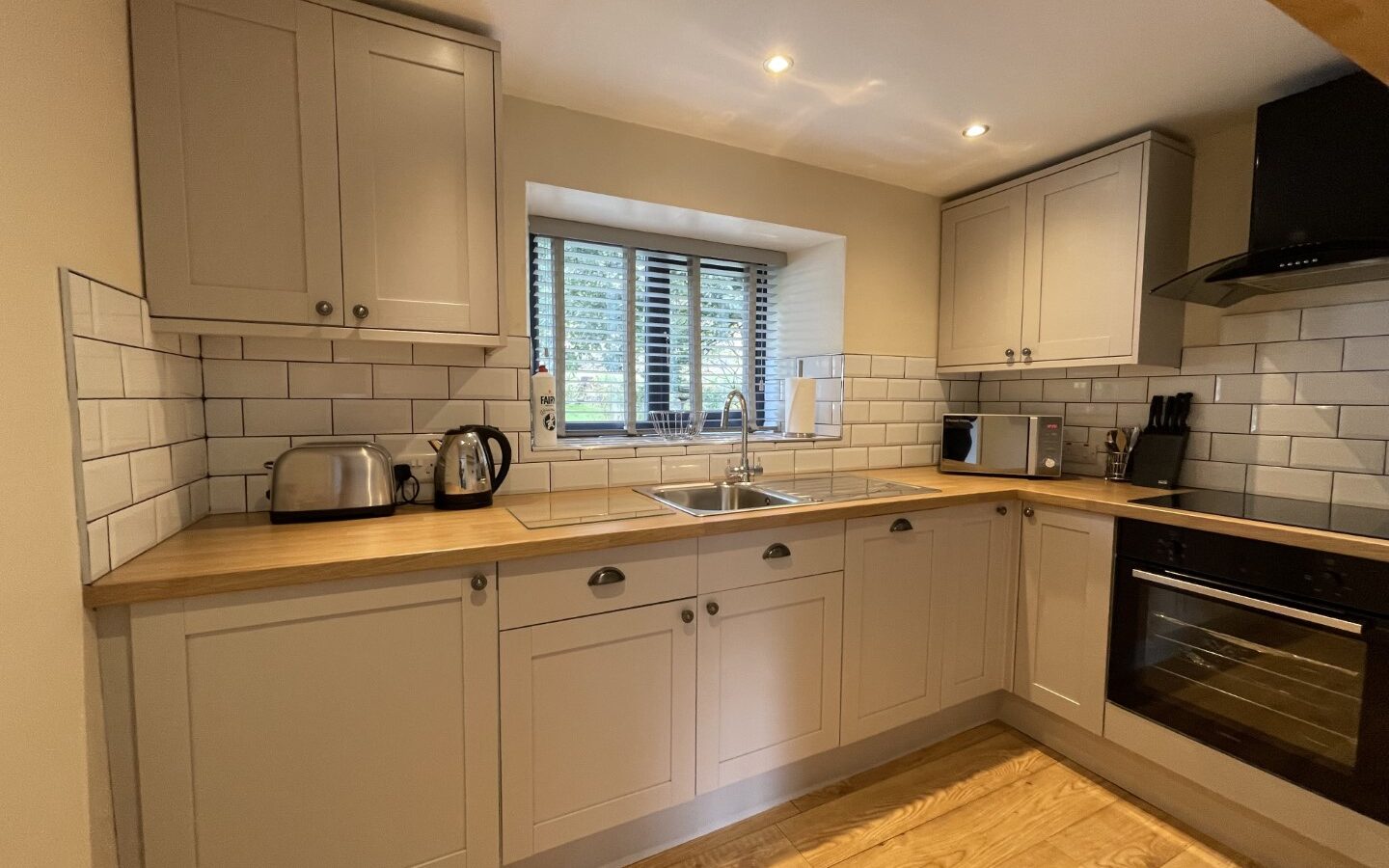 A modern kitchen nestled in Trallwm Cottages, featuring wooden countertops, white cabinets, a stainless steel sink, toaster, kettle, and a built-in oven. A serene blend of contemporary design amidst the tranquility of Forest Cottages.