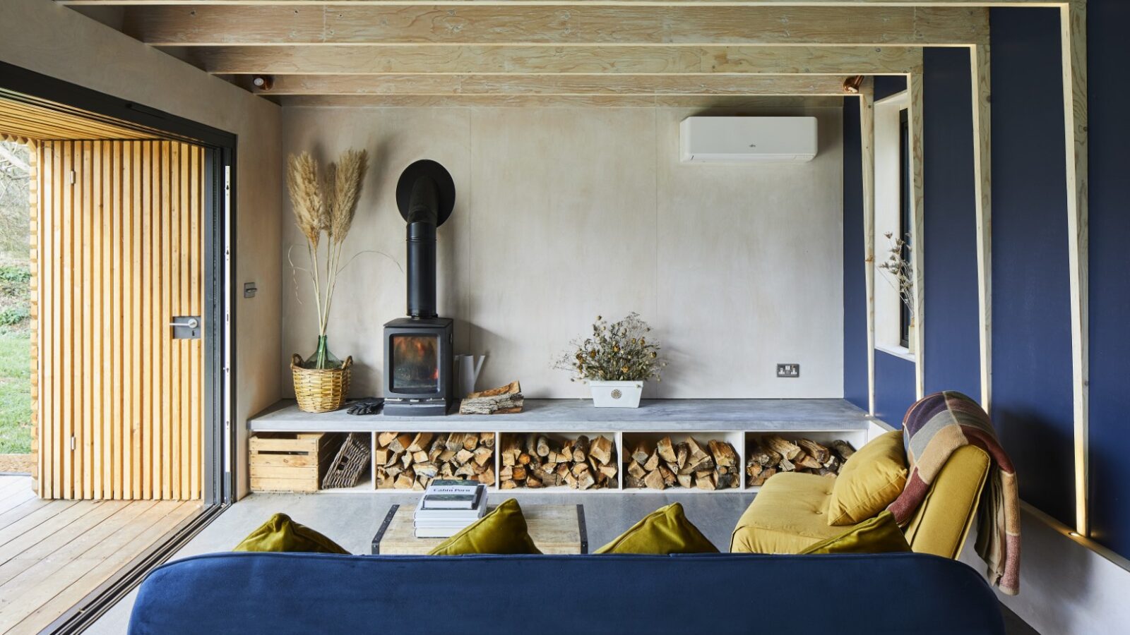 Modern living room with a wood-burning stove, stacked firewood, a blue sofa, and a large open doorway with wooden panels, offering the perfect retreat for Secret Escapes.