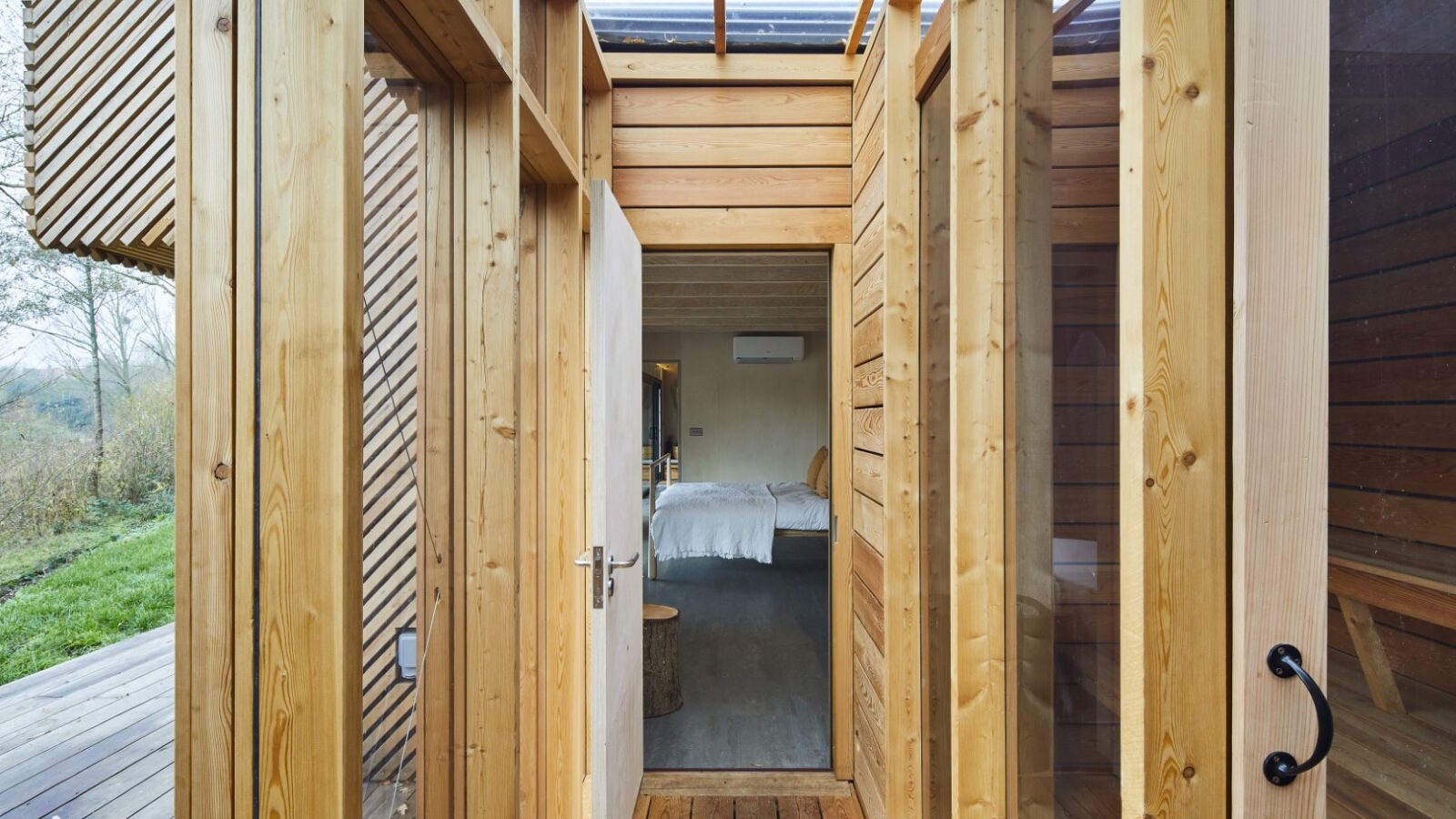 A wooden hallway, reminiscent of Browning Bros' rustic style, leads to a bedroom with a double bed, all visible through a glass door. The walls boast vertical slats and inviting windows, making it feel like your own Secret Escape.