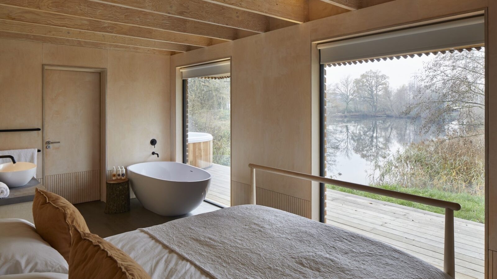 A minimalist bedroom crafted by Browning Bros features wooden walls, a large bed, and a freestanding bathtub beside a window overlooking the serene lake scene—a true Secret Escape.