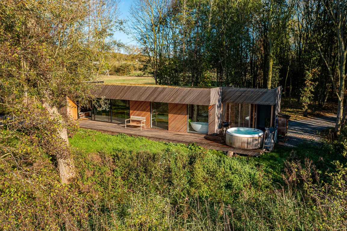 A modern, rectangular cabin by Browning Bros features a wood exterior, large windows, and a hot tub on a wooden deck, surrounded by trees—a true Secret Escape.