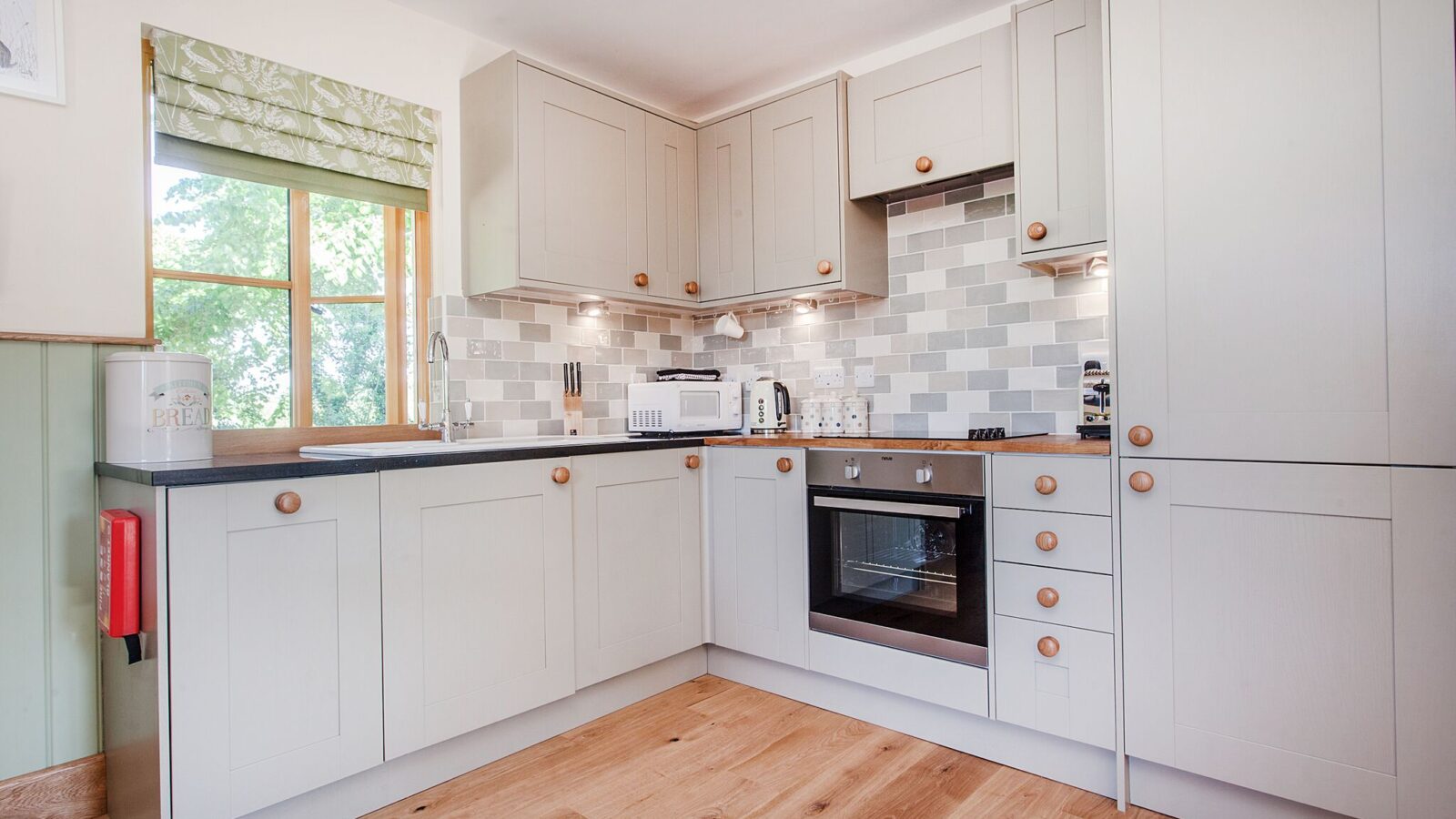 Modern kitchen with light gray cabinets, wooden handles, and a patterned backsplash reminiscent of the cozy vibe of Lavender Hill Treehouses. Anchored by a wooden floor and appliances thoughtfully placed near a window, it brings a touch of natural charm to urban living.