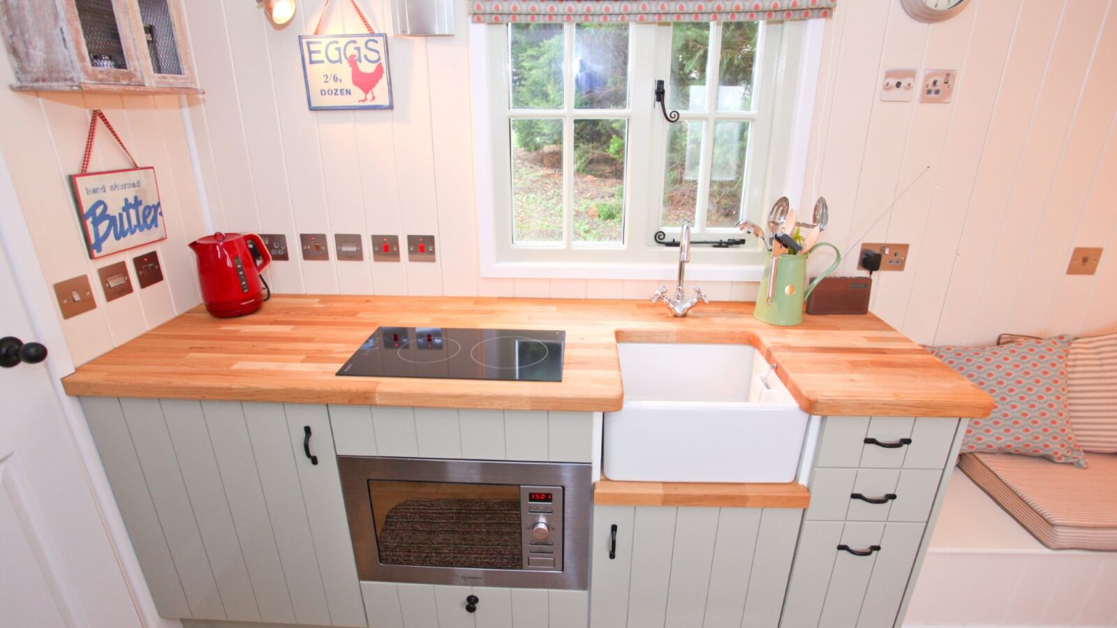 A cozy vintage-style kitchen at Abbey Farm features a wooden countertop, a small sink, and a built-in stove. A red kettle, microwave, and sign decor grace the walls. A patterned curtain adorns the window, while a cushioned bench cozily sits in the corner.