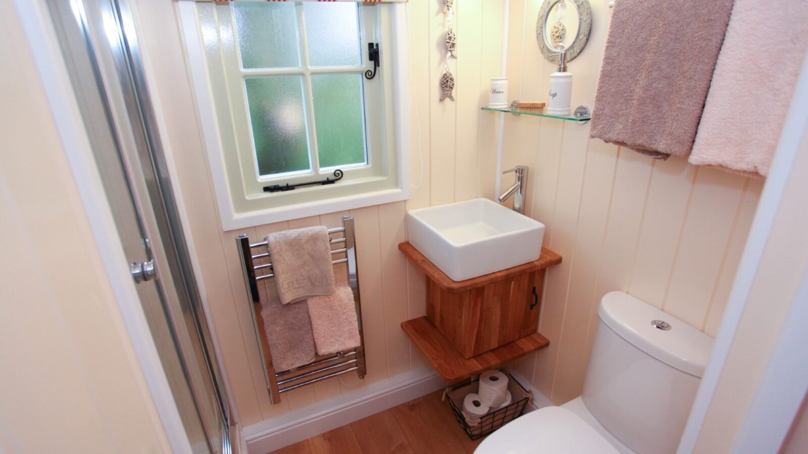 A small, cozy bathroom reminiscent of a Shepherd's Hut, with a window dressed in striped curtains. It features a wooden sink vanity, a towel rack with folded towels, a toilet with ample paper stocked, and a glass shower door. The walls are light-colored with vertical paneling.