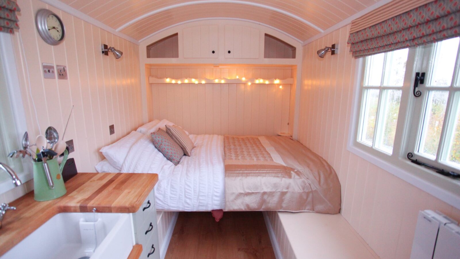 Cozy tiny house interior reminiscent of Shepherds Huts, featuring a neatly made bed with white and beige bedding, string lights above, and a window with patterned curtains. The compact kitchen area has wooden countertops and white cabinetry. The space is brightly lit and inviting.