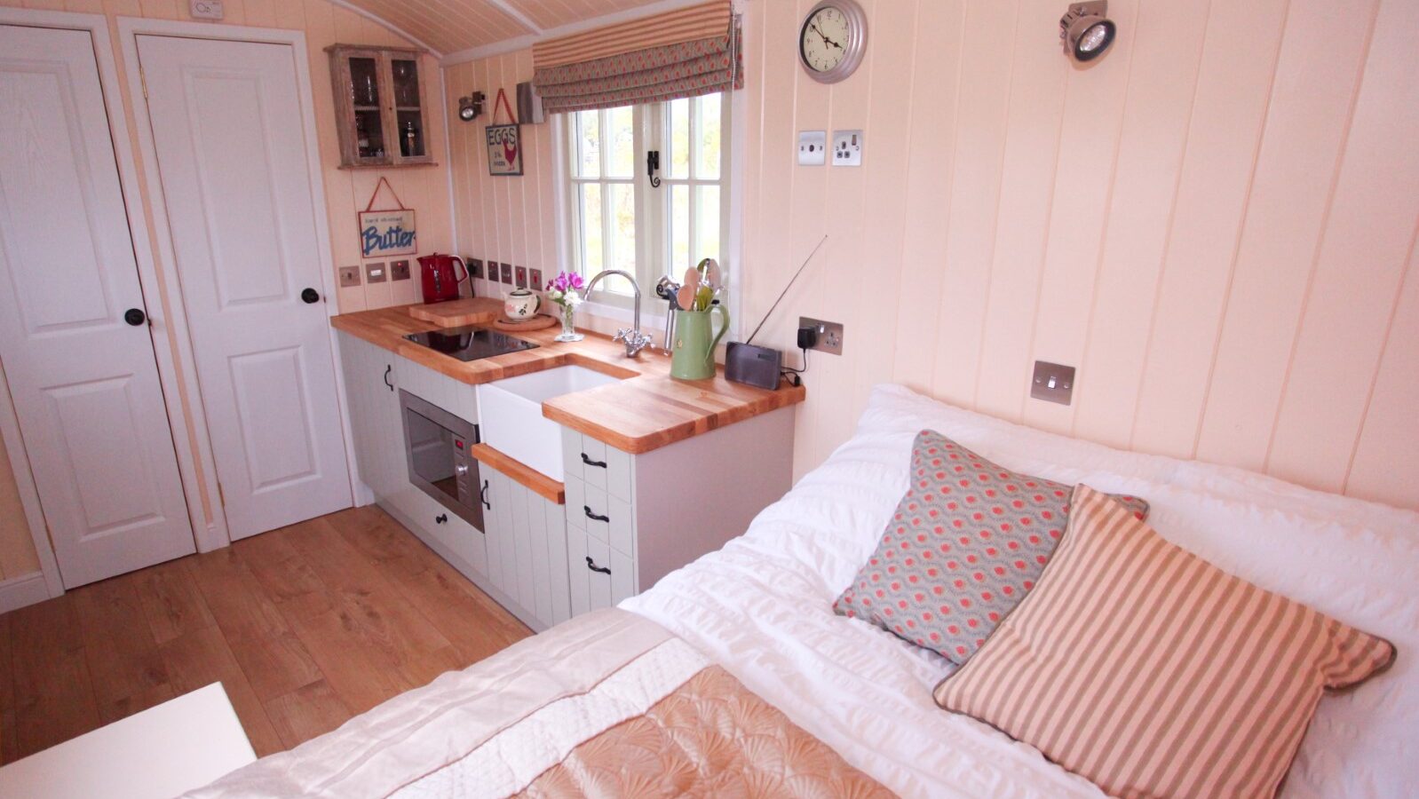 Cozy interior of a tiny house reminiscent of Shepherds Huts, featuring a bed with patterned pillows and a white quilt. The kitchen boasts wooden countertops, a small sink, and an oven. A window with blinds and decorative items adorn the walls, all enveloped in light wood paneling for charm.