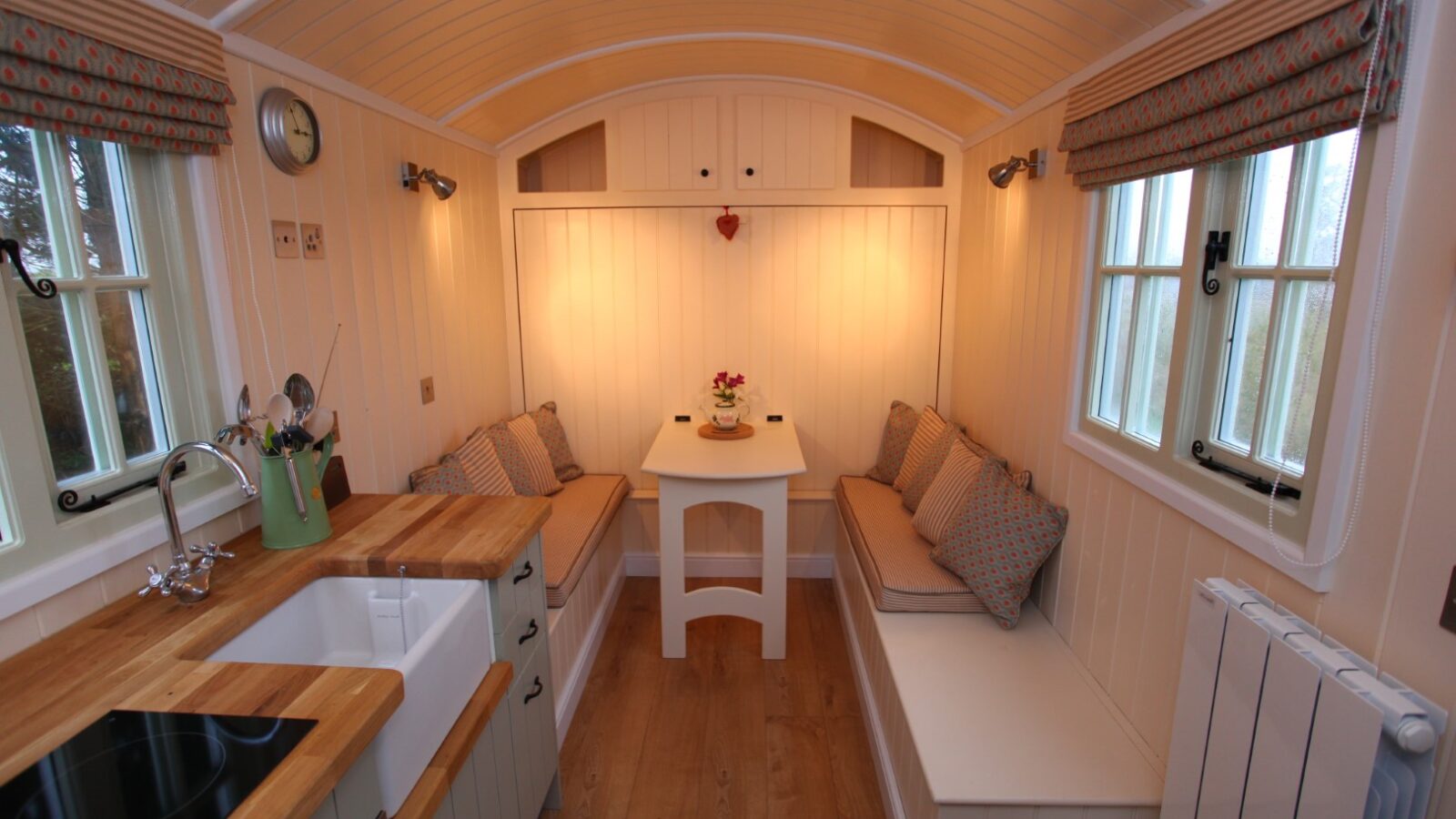 A cozy wooden interior in a Shepherds Hut features a small kitchen with a sink, stovetop, and counter on the left. A dining table with a flower pot, flanked by cushioned benches, rests in front of a softly lit wall. Windows with patterned curtains line the right side.