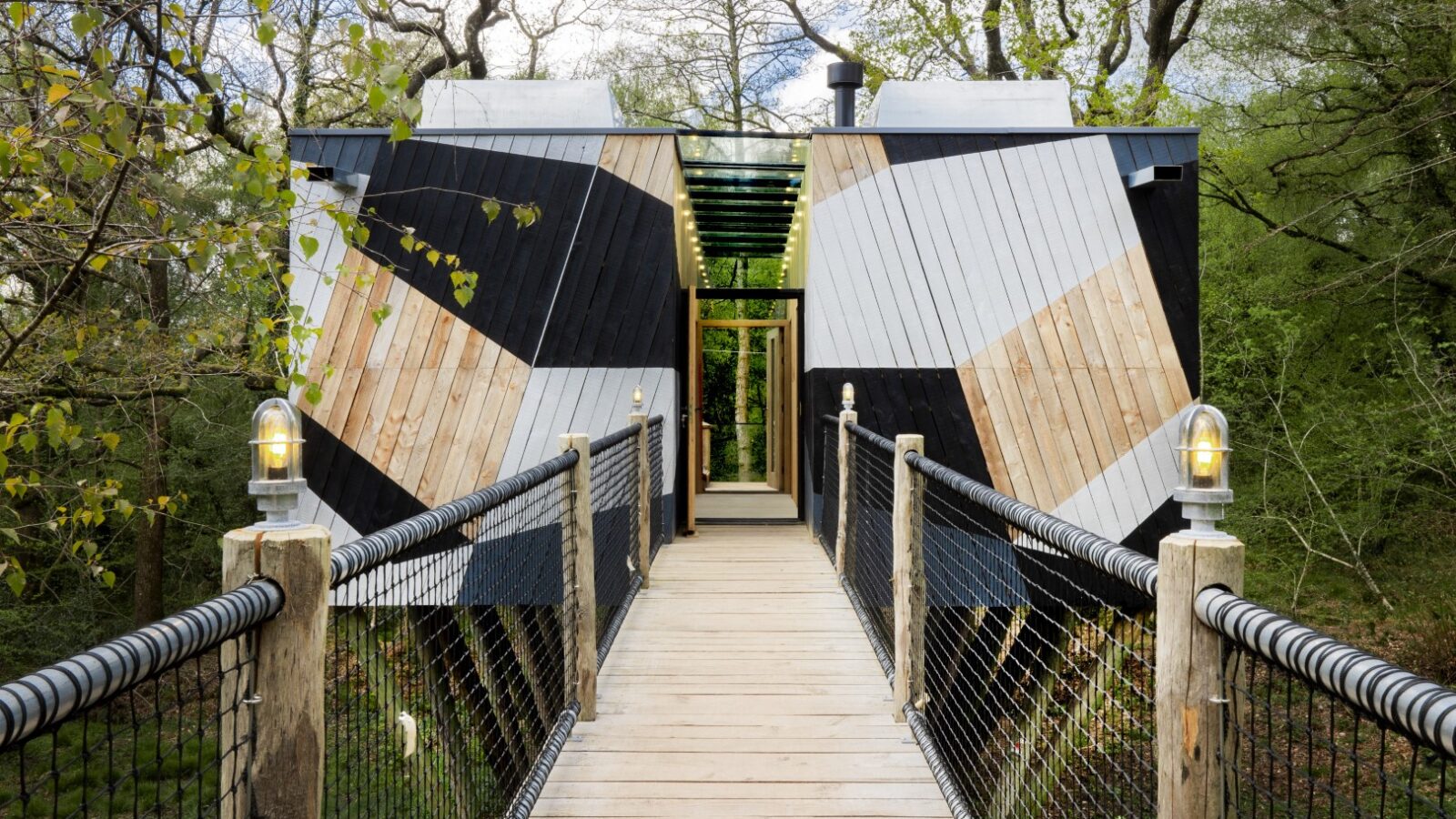 The Dazzle Treehouse is a modern geometric retreat, boasting a charming bridge amidst the trees and showcasing varied wood patterns on its exterior.