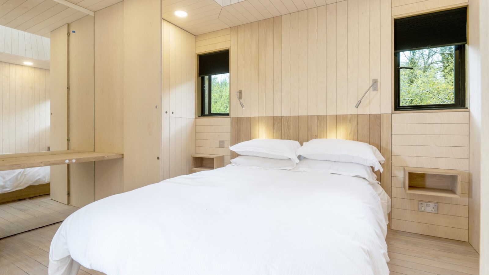 The Dazzle Treehouse offers a modern bedroom with a wooden interior, featuring a double bed adorned in crisp white bedding. Twin windows invite natural light, while built-in wall lights add subtle illumination to this serene space.