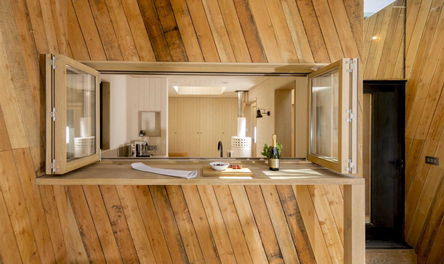 Open wooden window shutters on a diagonal wood-paneled wall reveal an interior kitchen with countertops and a cutting board, evoking the charm of a Dazzle Treehouse nestled in nature.