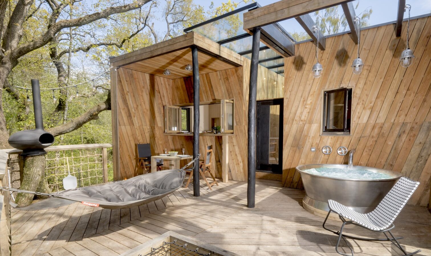 Discover the Dazzle Treehouse, a charming wooden cabin with a patio featuring a hammock, outdoor table, modern bathtub, and lounge chair—all nestled among the trees.