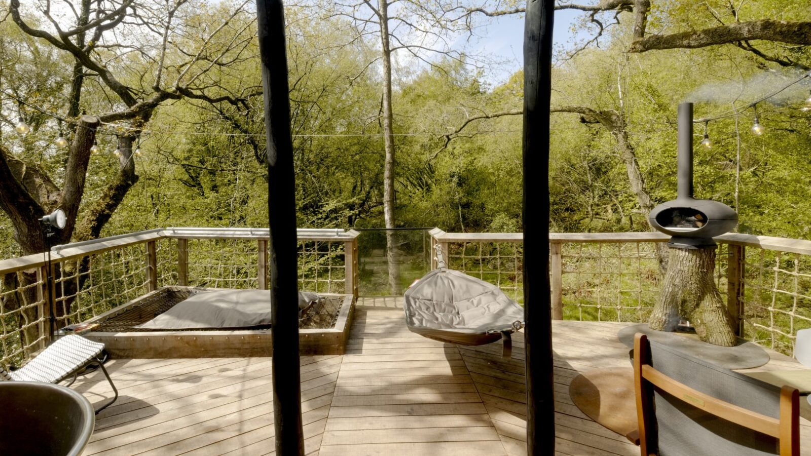 The Dazzle Treehouse features a wooden deck with chairs, a hammock, a small square sandpit, and a stove, all nestled among leafy green trees.