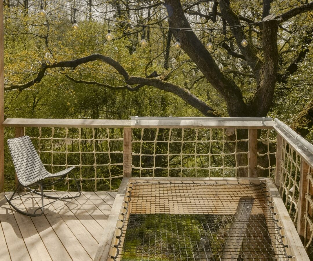 A wooden deck with a netted section and a woven chair overlooks the leafy trees of the dazzling Dazzle Treehouse forest.