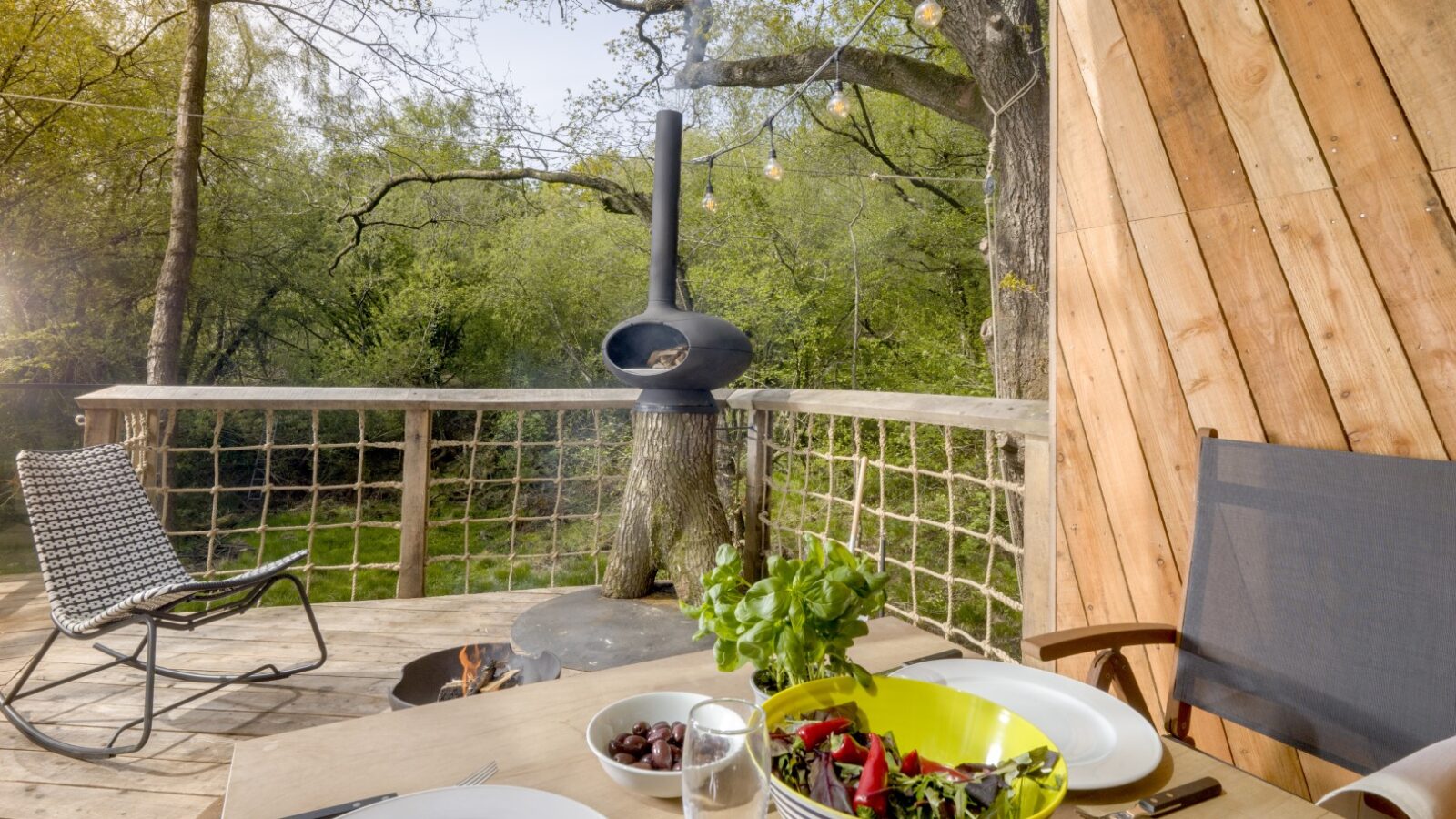 A cozy treehouse deck awaits, with a table set for two that dazzles with a spread of salad and olives. Nearby, a chair invites relaxation by the warm glow of a wood-burning stove, enveloped by a serene canopy of trees.