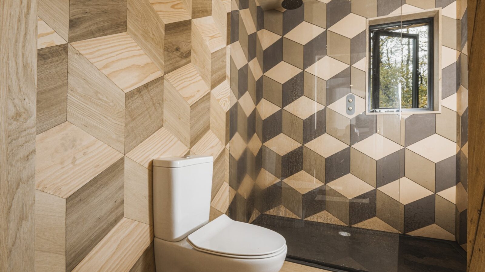 Step into the Dazzle Treehouse's modern bathroom, where a geometric tile design captivates. A pristine white toilet complements the shower area, illuminated by natural light streaming through a window.