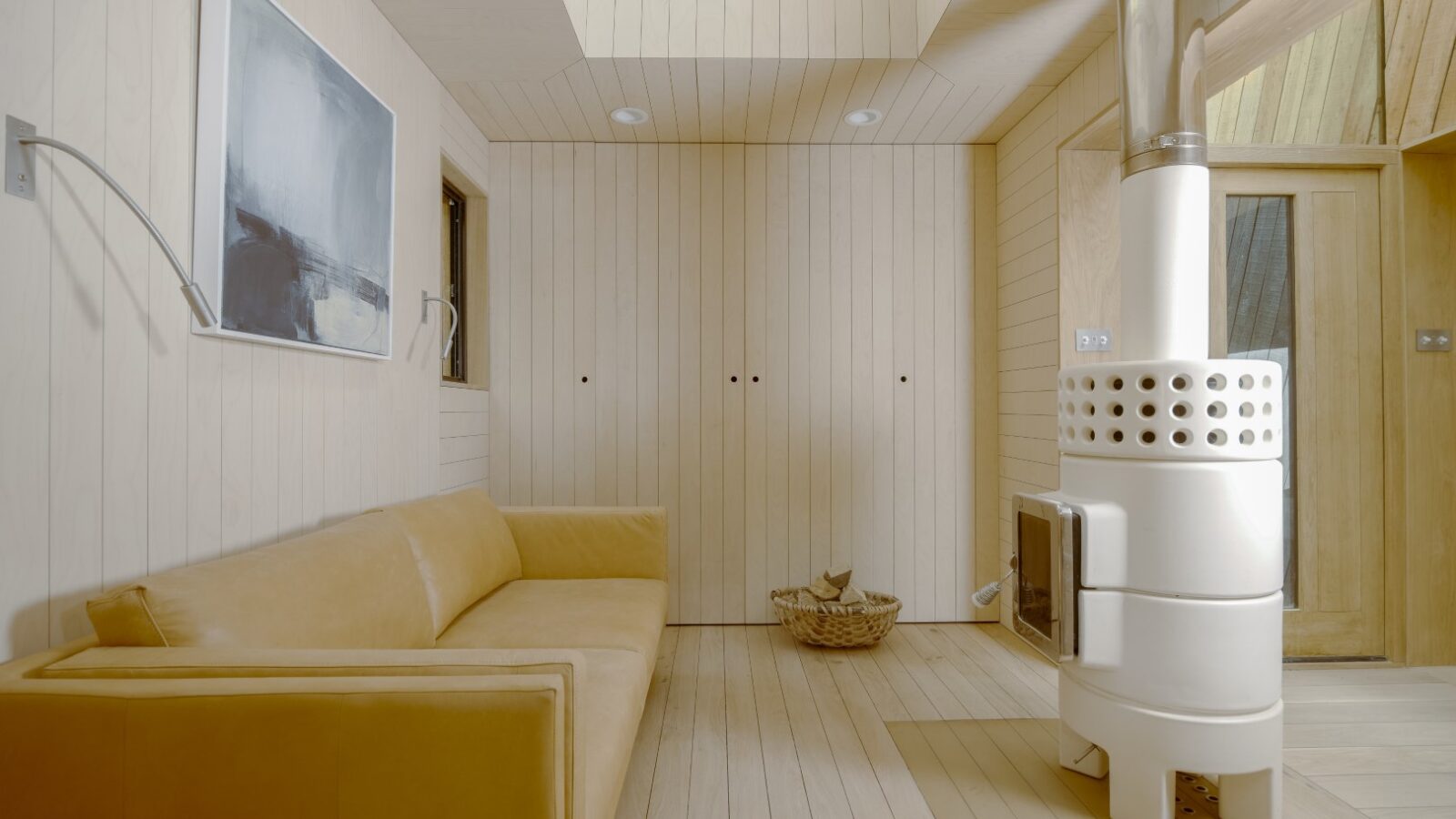 Experience the charm of a minimalist living room in the Dazzle Treehouse with its beige couch, contemporary wall art, and a modern cylindrical wood stove. A basket accents this serene space, perfectly blending simplicity with the dazzle of nature's retreat.