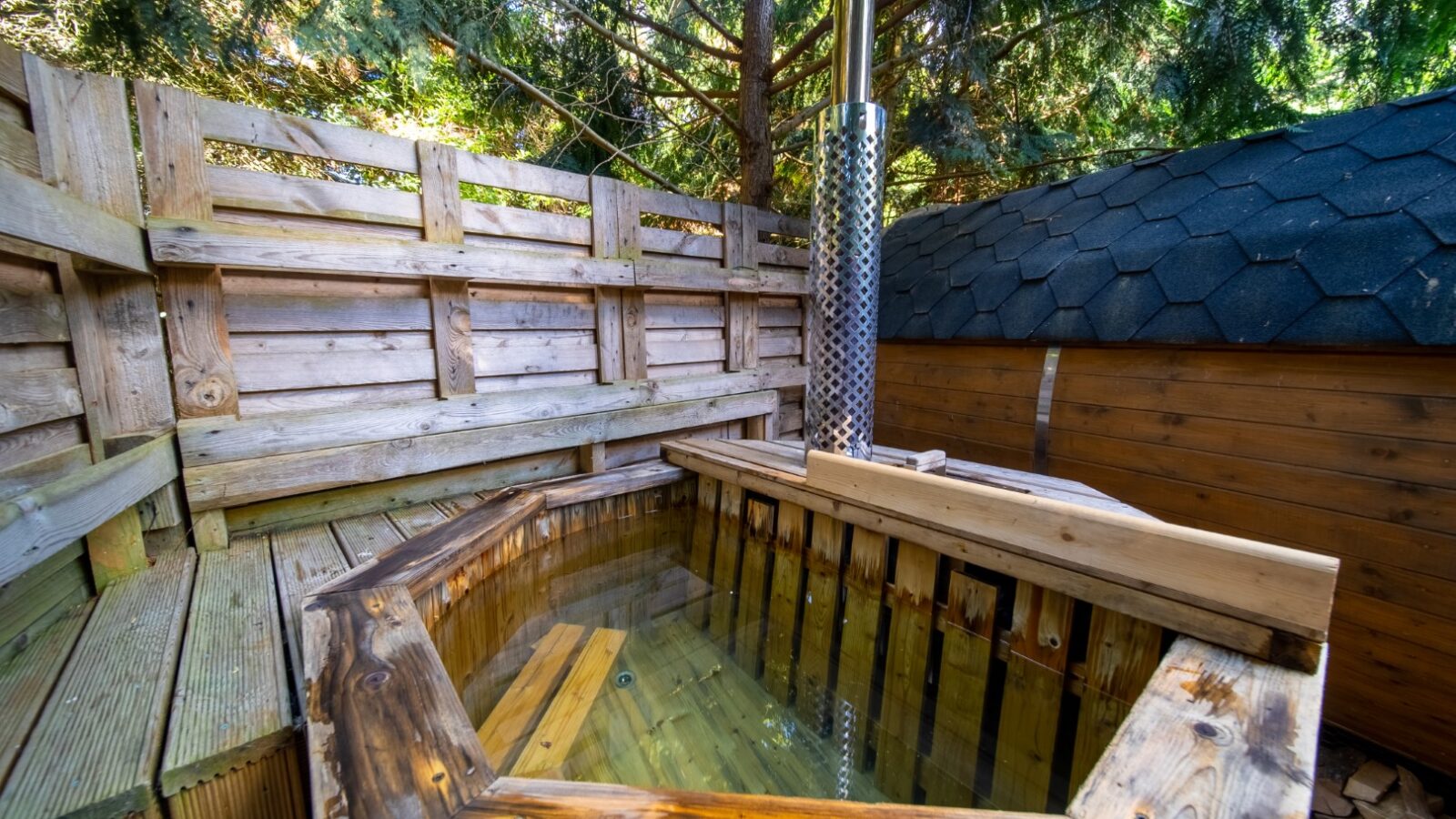 A wooden outdoor hot tub filled with clear water, surrounded by a wooden deck and a fence, offers a tranquil and rustic Lahtle Wood experience. Tall evergreen trees provide shade, while a wood-burning stove is attached to the side of the tub. The perfect setting for glamping in nature's embrace.