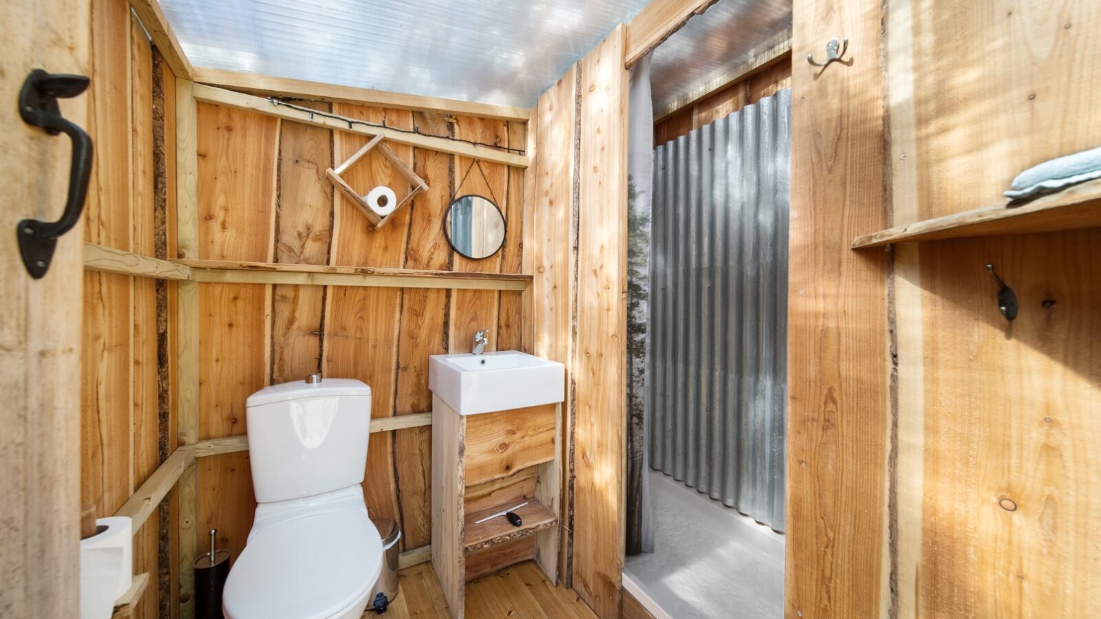 A glamping experience with a rustic wooden bathroom featuring a white toilet and small sink. Above the sink, you'll find a round mirror and shelf with a toothbrush holder and towel. The translucent ceiling adds charm, while a door to the right leads to a shower area with corrugated metal walls.