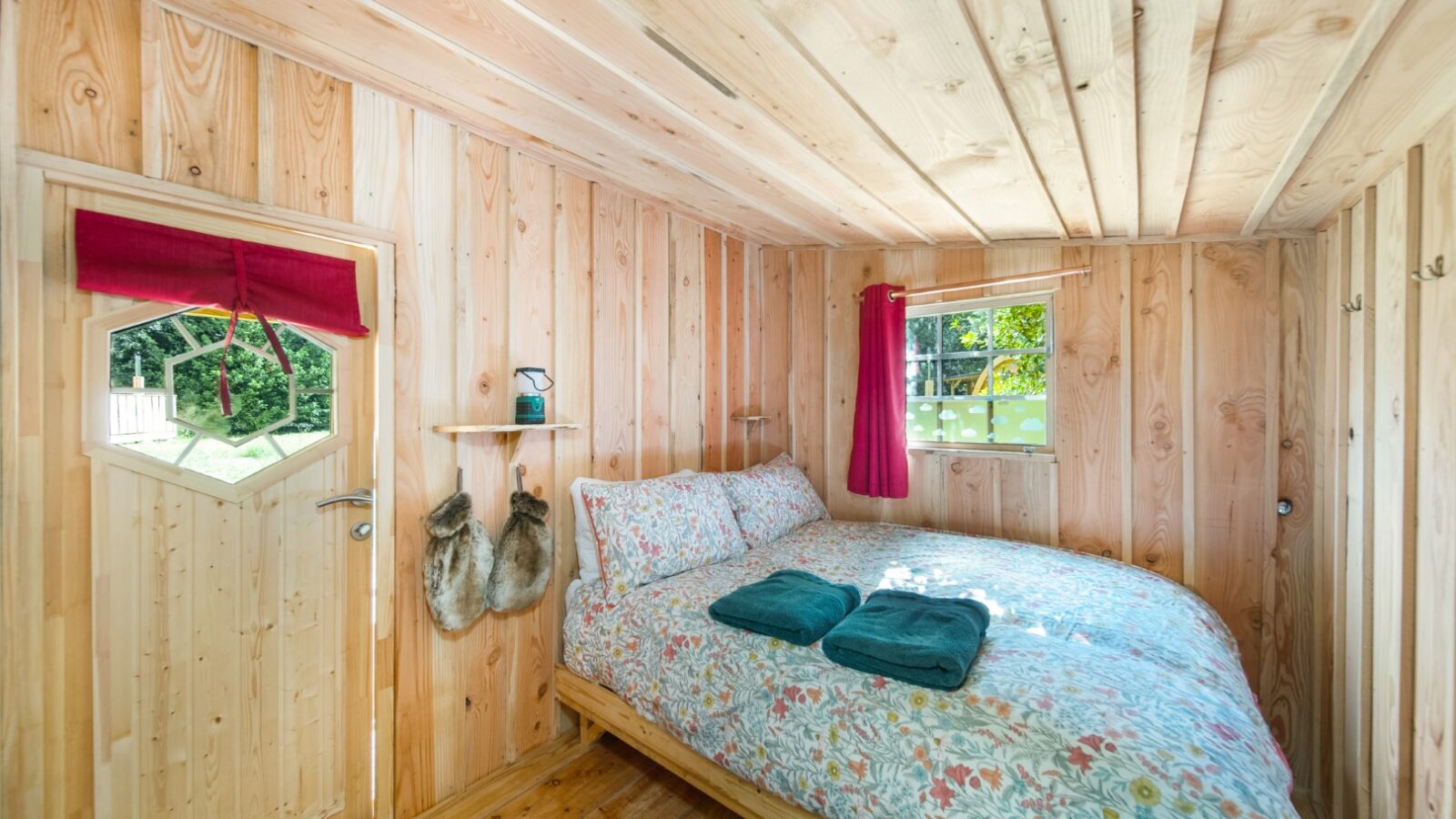 A cozy wooden room perfect for glamping with a double bed covered in floral bedding, two folded green towels, and a window with red curtains. A door with a hexagonal window features a red valance and two fur-covered slippers hanging beside it. Sunlight streams in through the window.