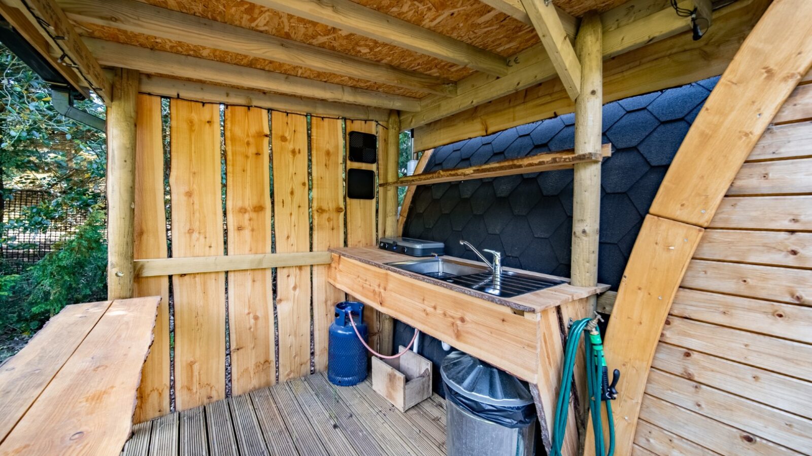 An outdoor wooden kitchen with a sink and countertop. A blue gas tank and a green hose are under the sink. The walls and ceiling are made of unfinished wooden planks, giving it a rustic feel perfect for glamping. The backdrop features a curved, shingled wall, and the floor is made of wooden decking.