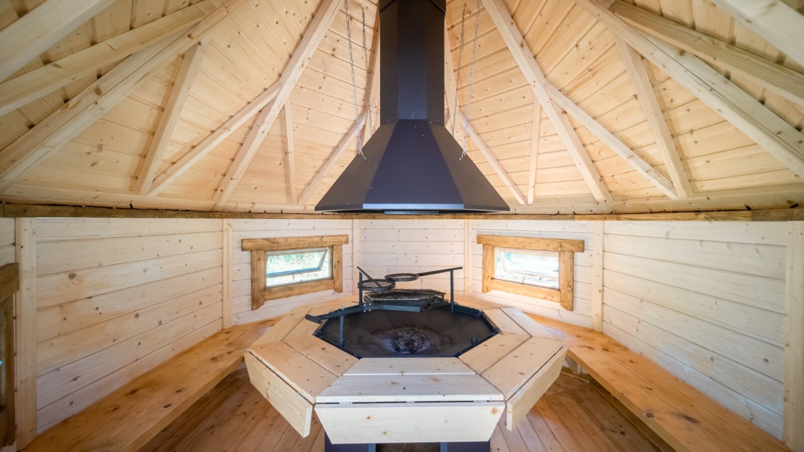 A cozy wooden cabin interior at Lahtle Wood features a central hexagonal grill surrounded by wooden benches. The walls and vaulted ceiling are made of light-colored wood, with three small windows letting in natural light, enhancing the warm ambiance of this glamping site.