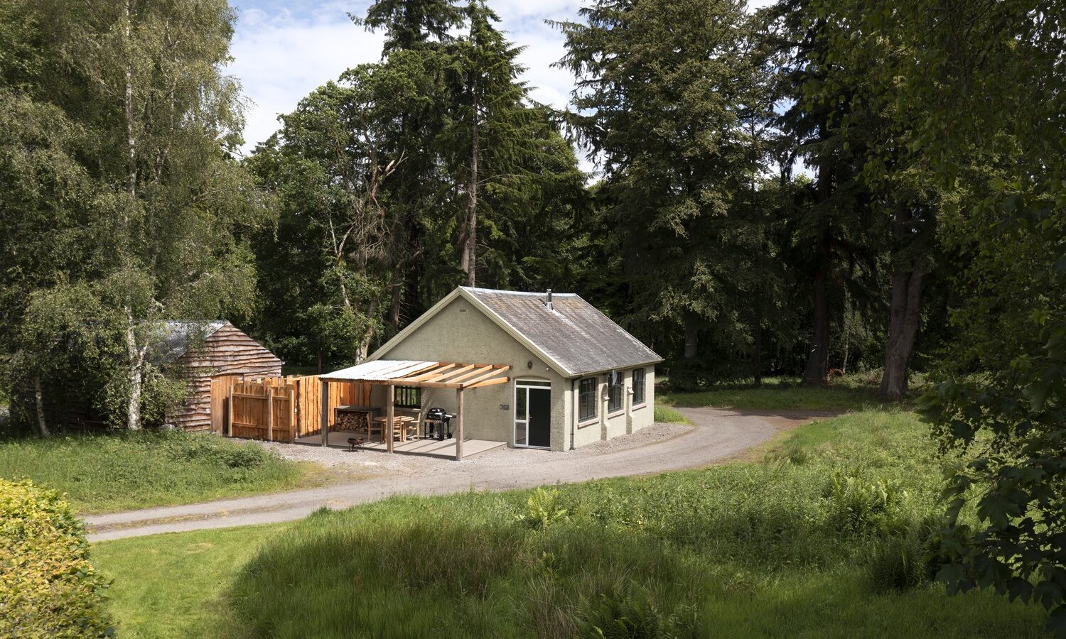 Nestled in a lush green forest, Keepers Bothy is a charming, single-story house with a wooden pergola. A gravel path leads to its entrance, while towering trees embrace the home, providing serene and natural surroundings.