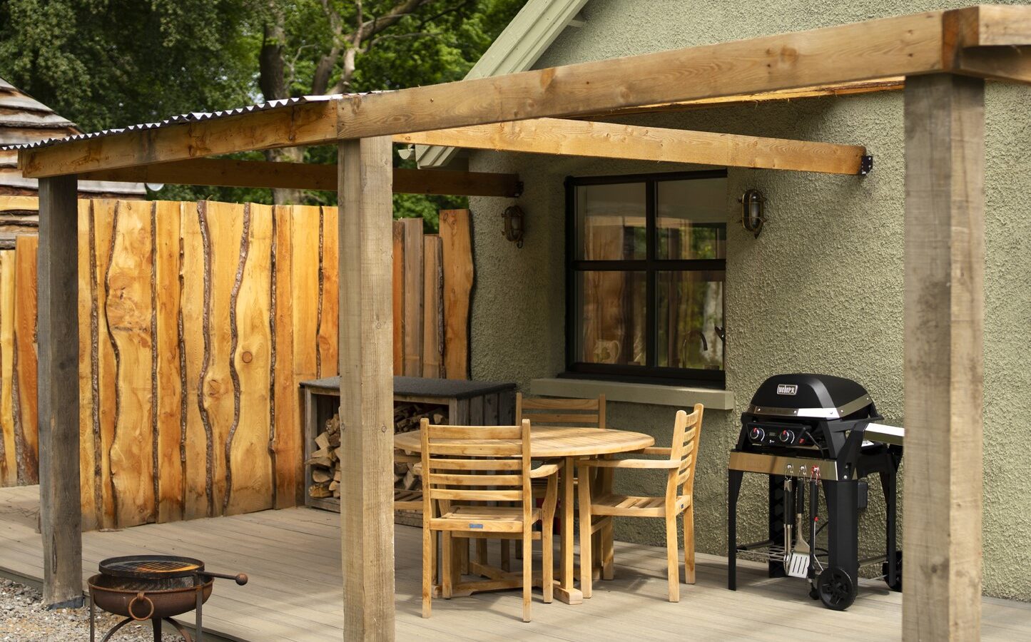 The wooden deck at Keepers Bothy features an outdoor dining set, a barbecue grill, and a fire pit, all nestled beside the rustic house with its charming wooden fence.