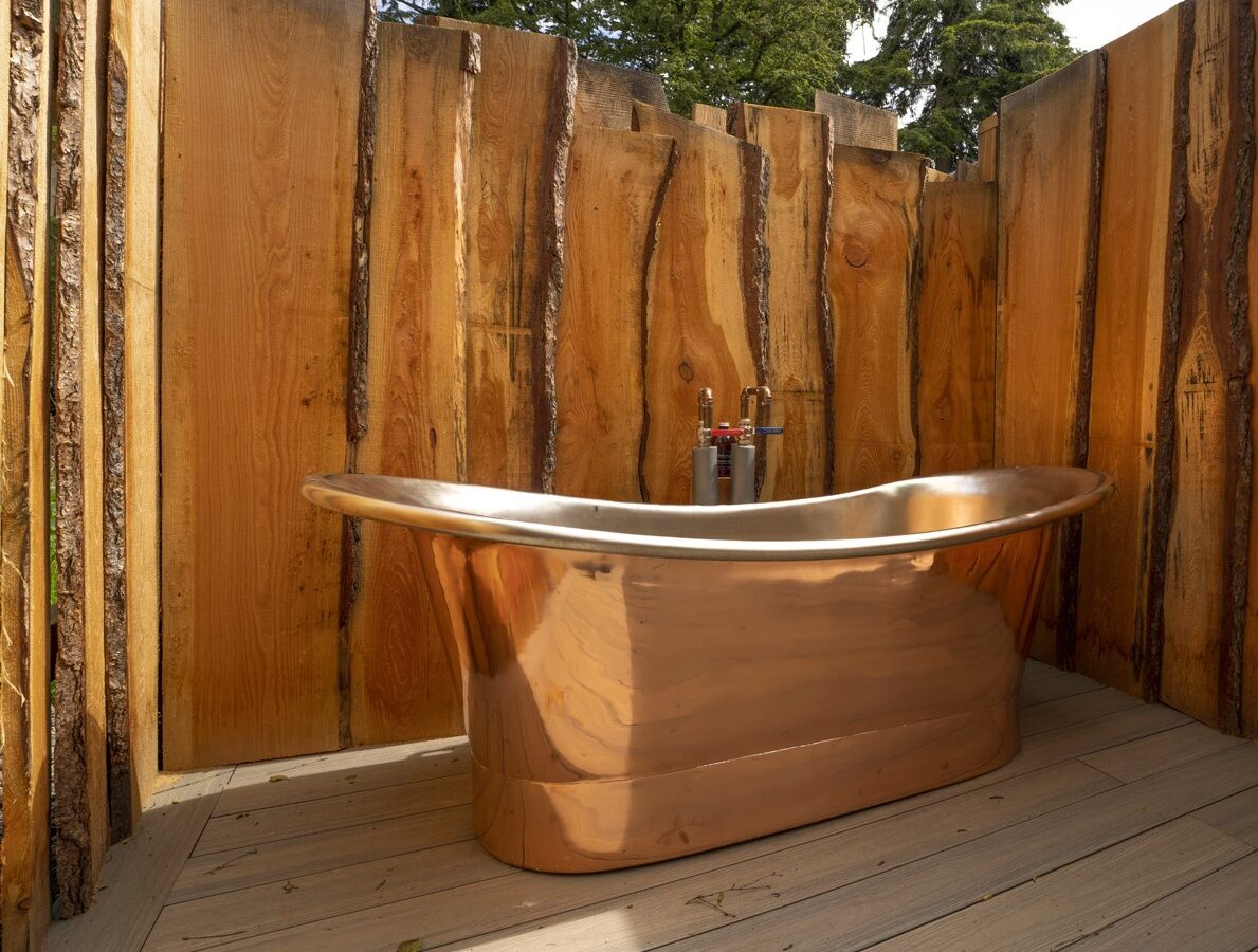 A luxurious copper bathtub is placed outdoors at the Keepers Bothy, surrounded by rustic wooden walls. The area is set against a backdrop of lush green trees, creating a serene and natural bathing environment.