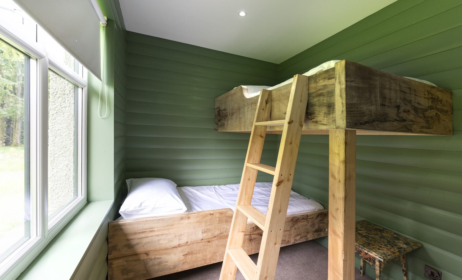 A small, bright room with green wooden walls reminiscent of a cozy Keepers Bothy features a wooden bunk bed. The lower bed is neatly made, and a ladder leads to the upper bed. A large window on the left lets in natural light, while a small stool sits near the corner.