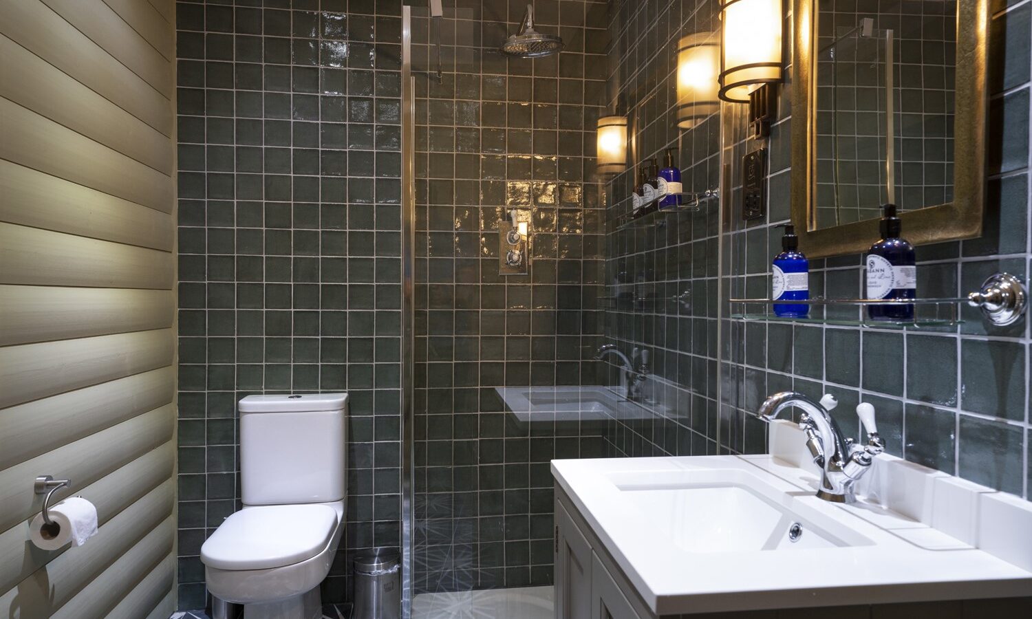 This modern rental bathroom features green tiled walls, a glass-enclosed shower, and sleek white fixtures with chrome accents. Illuminated by wall-mounted lights, the space includes a mirror above the sink. Perfect accommodation for those seeking style and comfort akin to Keepers Bothy.