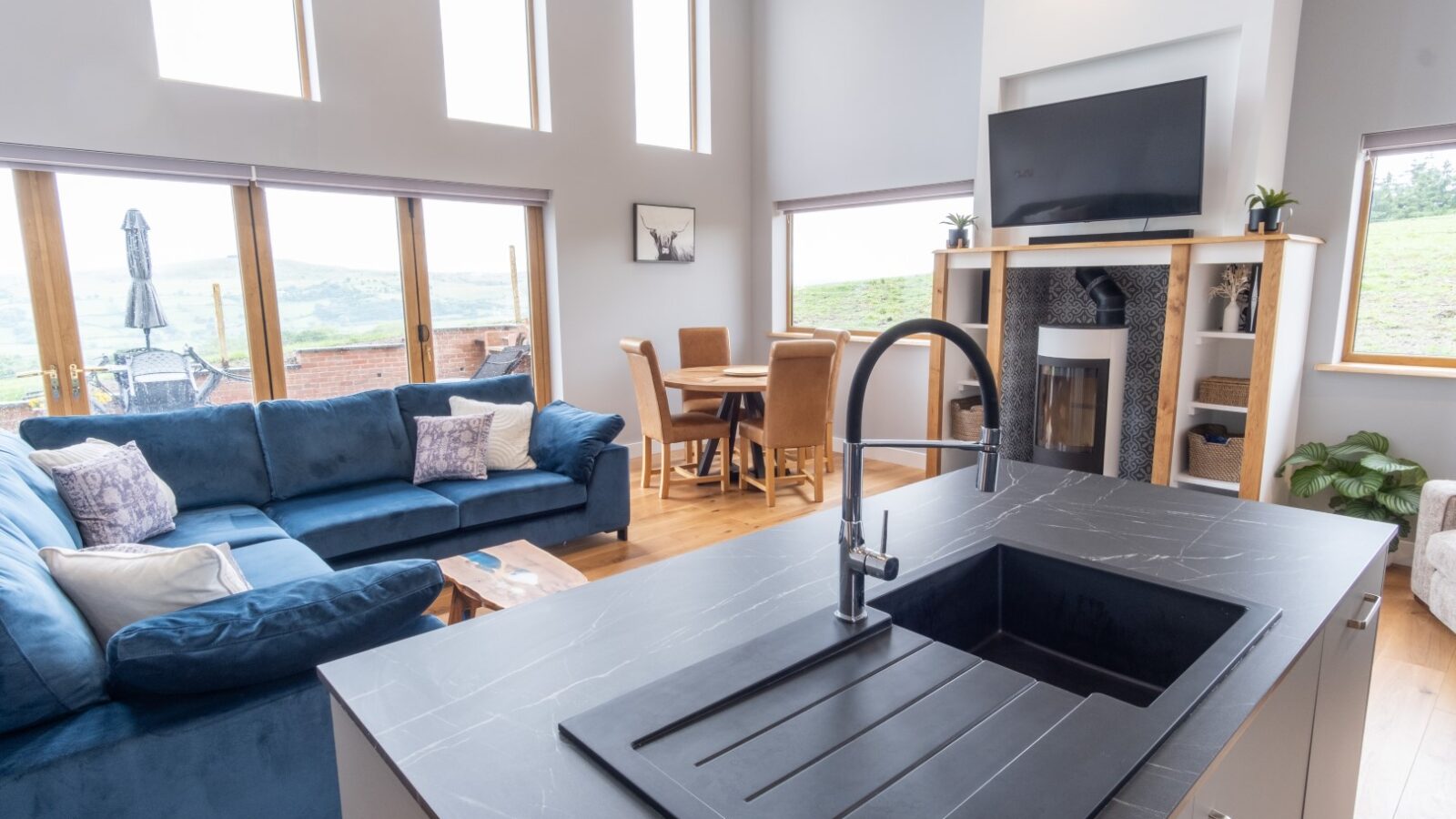 Discover the charm of this modern living room at Lundy Lodges, featuring a blue sofa, dining table, large windows, black countertop island, and wall-mounted TV. Whether you're a travel enthusiast or seeking cozy lodging, this space offers the perfect blend of comfort and style.