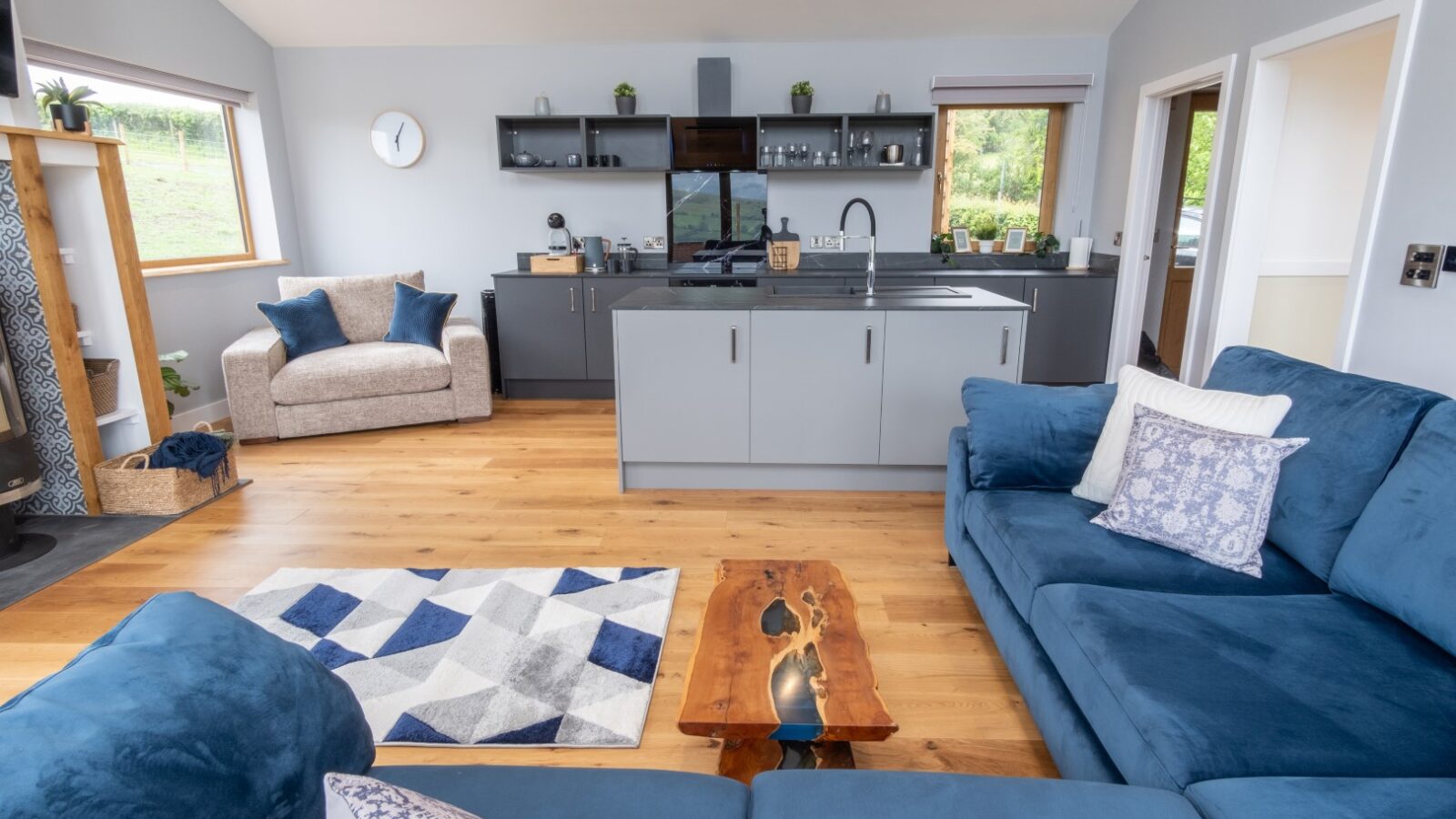 The Lundy-inspired open-concept living area boasts blue sofas and elegant wood flooring, seamlessly flowing into a modern kitchen with grey cabinetry and sleek black countertops, perfect for upscale lodges.