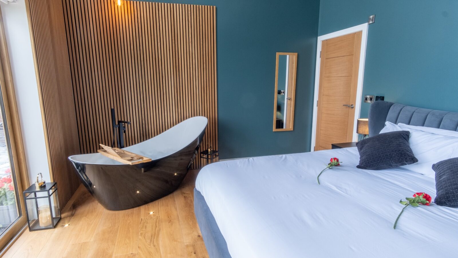 A modern bedroom at Lundy Lodges features a freestanding black bathtub, a large bed adorned with pillows and roses, a vertical wood accent wall, and an elegant mirror.