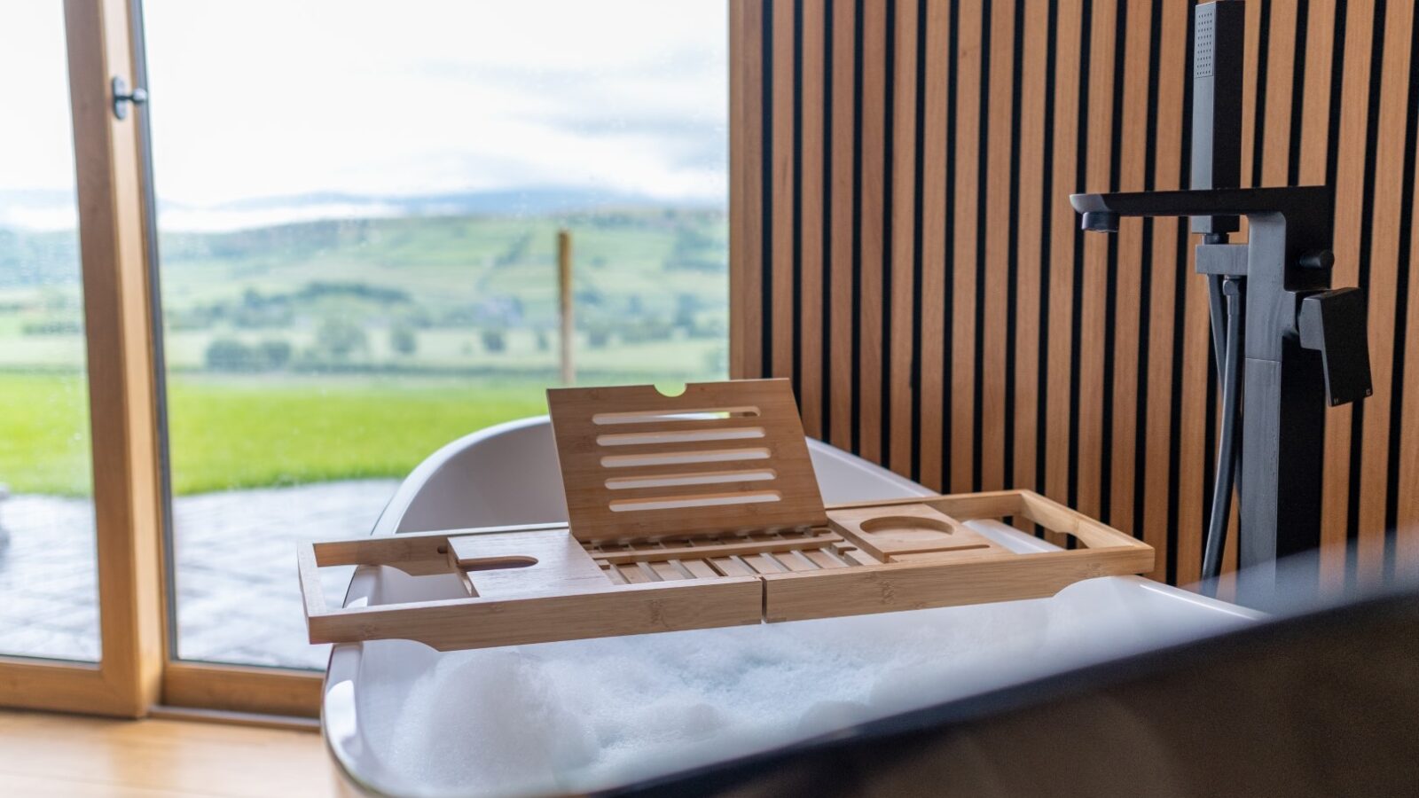 A modern bathtub with a bamboo tray, filled with bubbles, sits invitingly near a large window at Lundy Lodges, offering a breathtaking view of the scenic countryside.