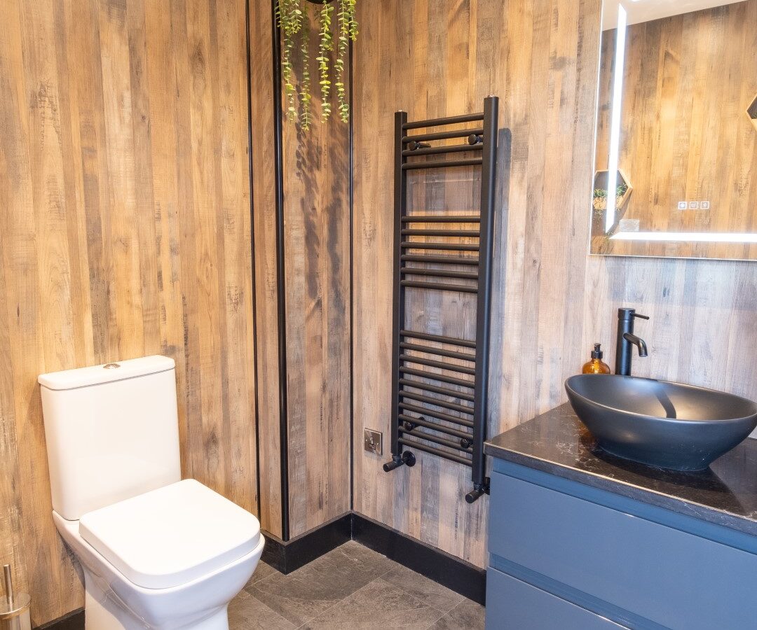 This cozy Lundy bathroom features wood panel walls, a white toilet, a black towel rack, a vanity with a sleek black bowl sink, and a small hanging plant—a perfect retreat reminiscent of rustic lodges.