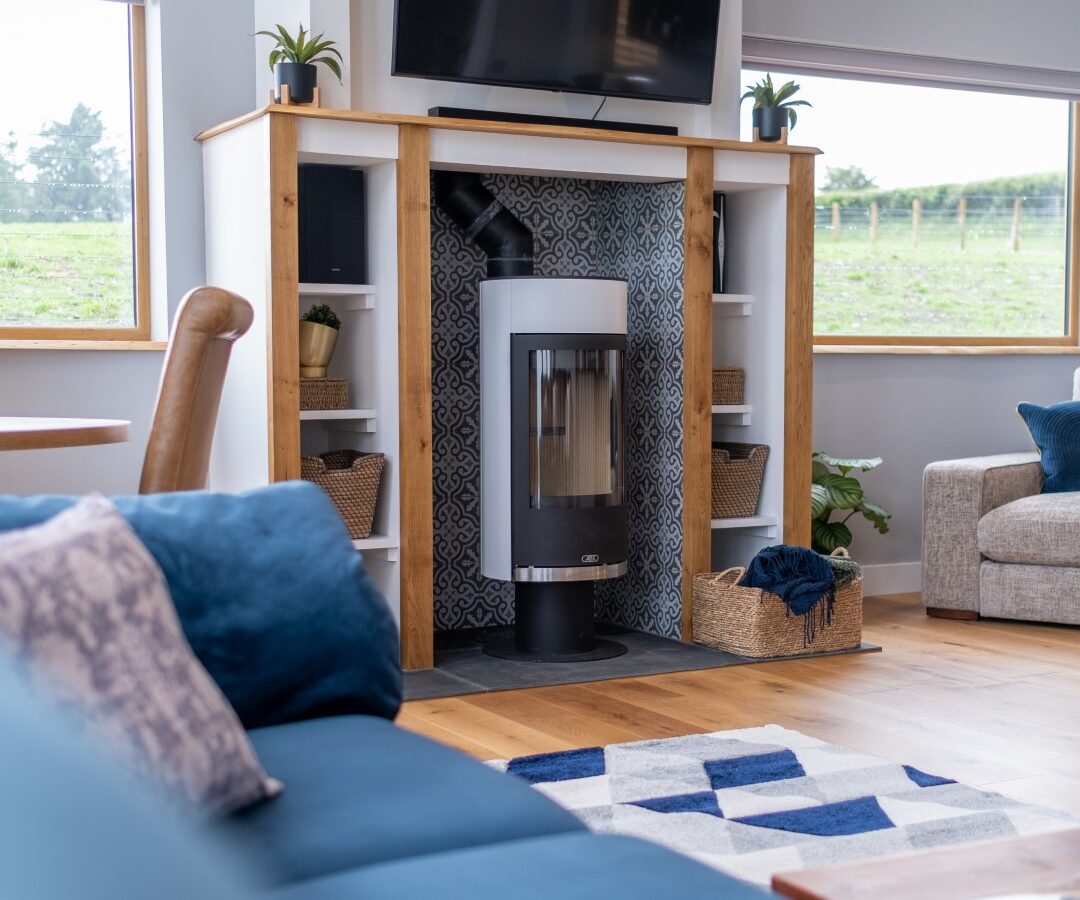 A modern living room in Lundy Lodges features a wood-burning stove with a television above it and blue seating. Large windows provide stunning views of greenery, making this the ideal accommodation for relaxation.