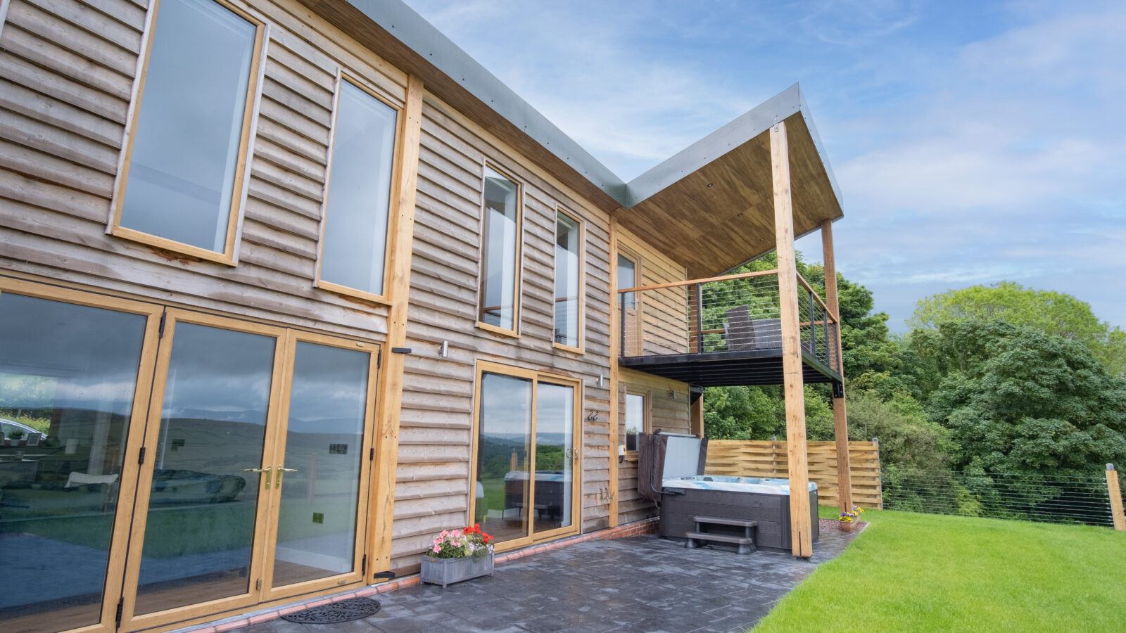 This modern two-story wooden house, part of the exquisite Lundy Lodges collection, features large glass windows, a balcony, and a patio. Nestled against a backdrop of trees and a lush lawn, it offers an idyllic retreat.