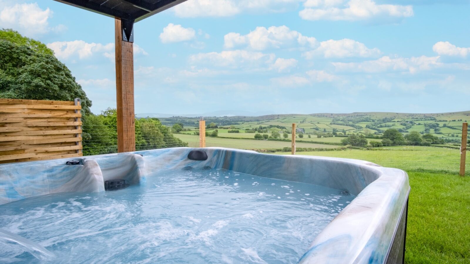 Enjoy the serene view of the countryside from a hot tub on a patio at our cozy Lundy lodges, with wooden fencing framing the clear blue sky and scattered clouds.