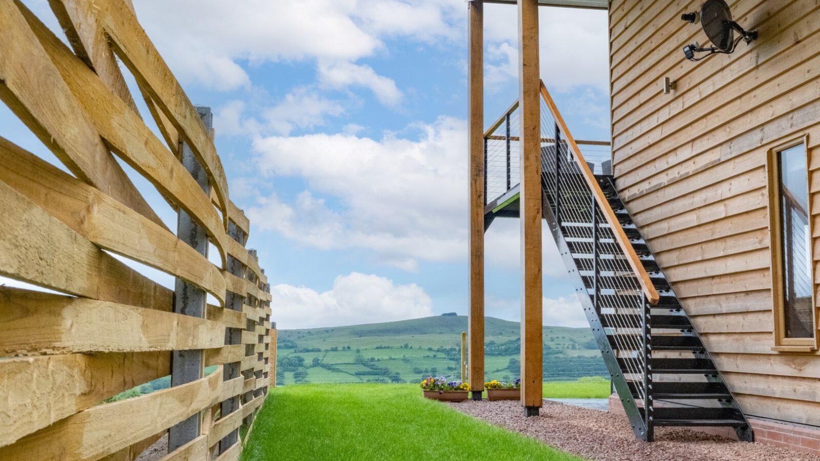 Nestled among lush green fields and rolling hills, this wooden-sided Lundy Lodges home features elegant metal stairs beside a charming wooden fence, all under a partly cloudy sky.