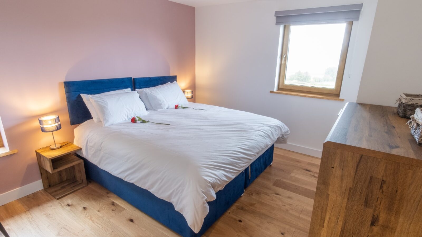 The Lundy bedroom boasts a large bed with a blue headboard and crisp white bedding. Lodges of style are echoed by two bedside tables with lamps, complemented by a wooden dresser and floor.