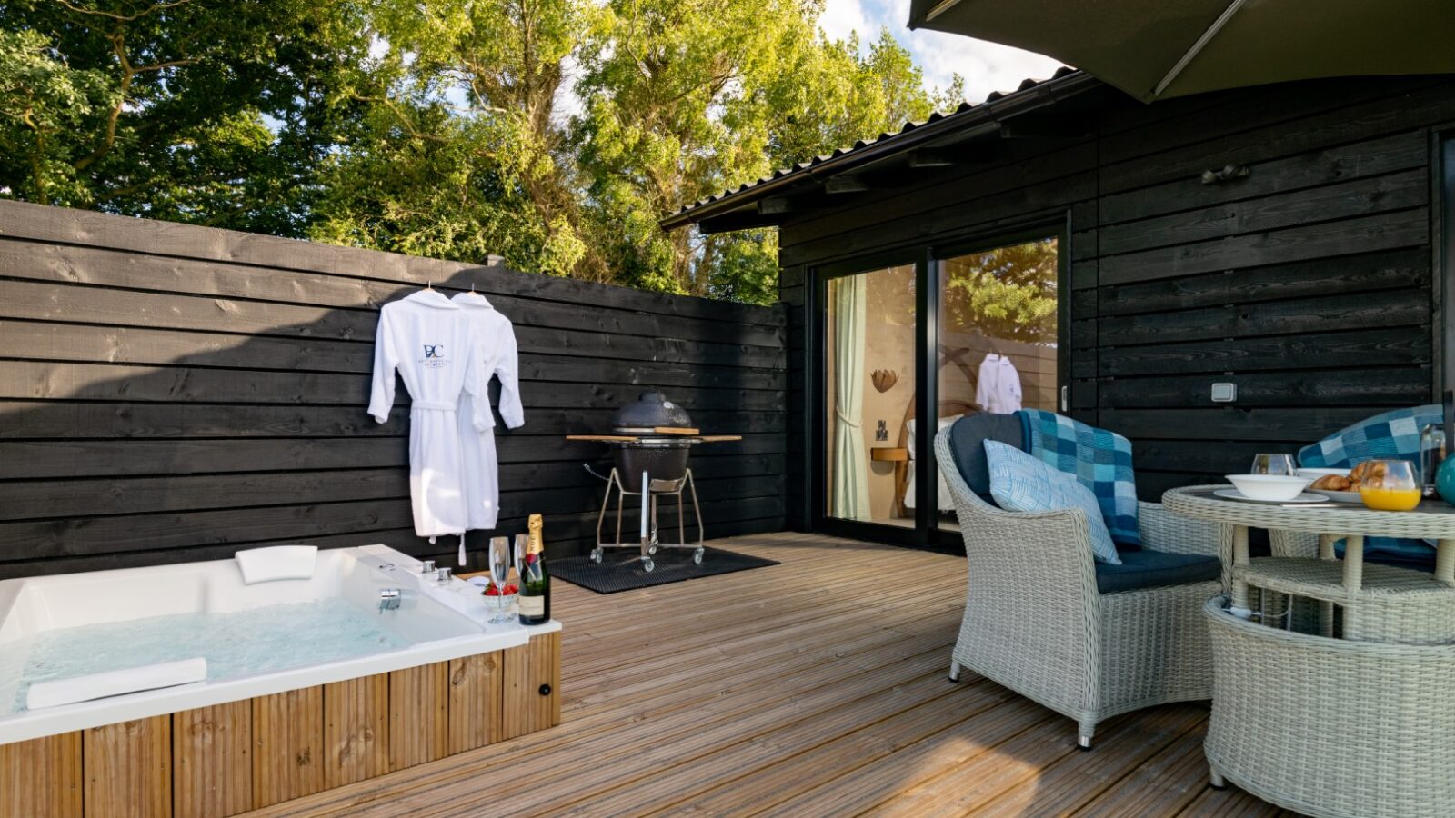 At Bettws Court Retreats, enjoy a wooden deck complete with a jacuzzi, wicker chairs around a table, a grill, and a bathrobe casually hanging on the black fence.