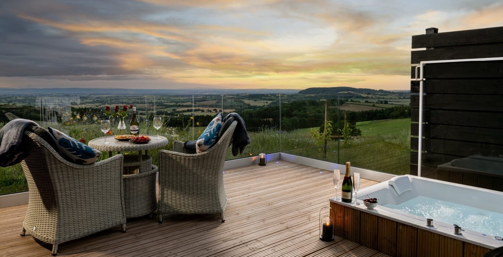 The outdoor deck at Bettws Court Retreats features wicker chairs and a table adorned with wine glasses, inviting guests to unwind. A hot tub adds a touch of luxury as you gaze over the expansive countryside at sunset.