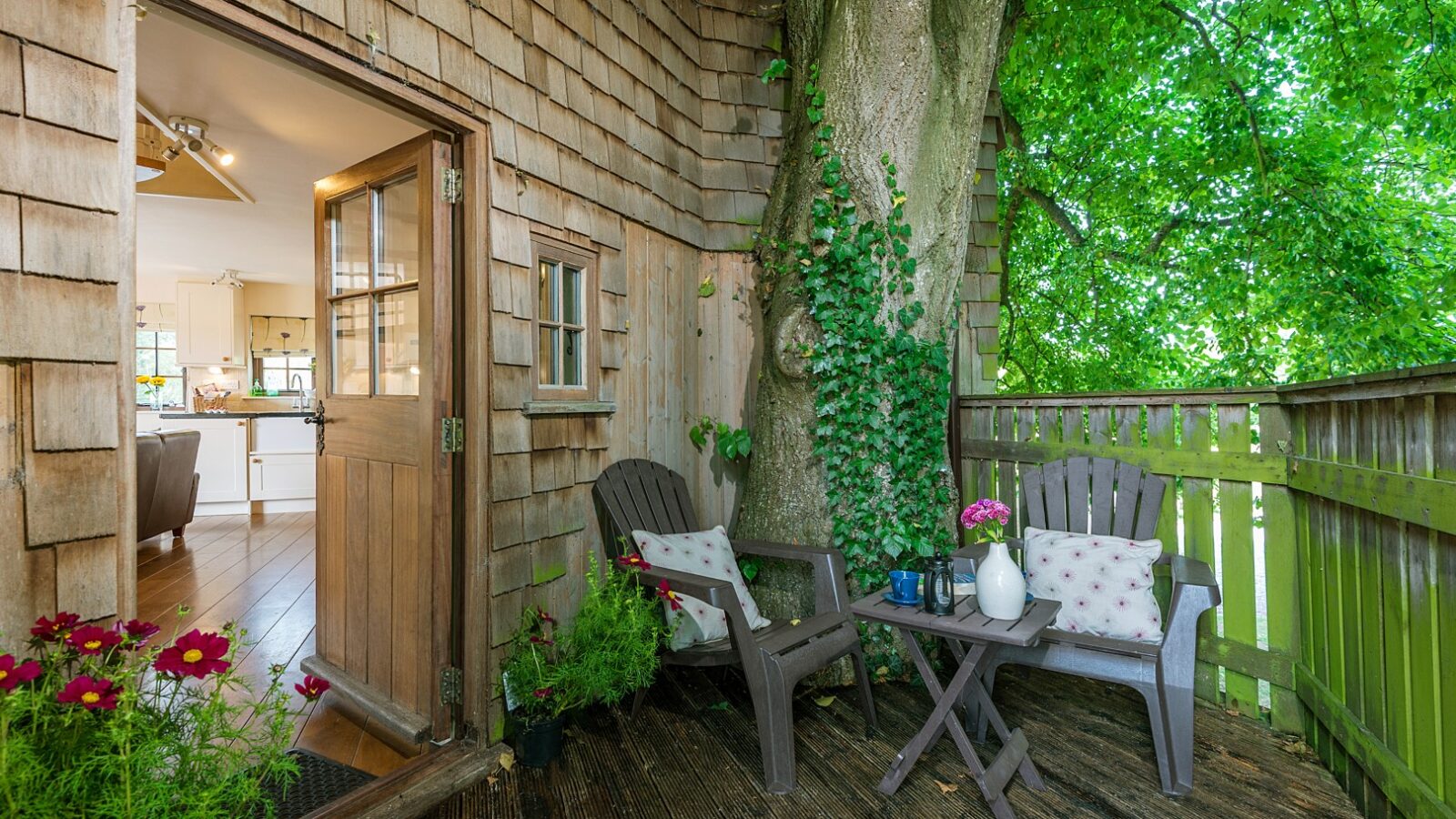 A wooden deck with two chairs and a small table, nestled among the greenery of Lavender Hill, leads into a cozy treehouse room with a light-colored interior.