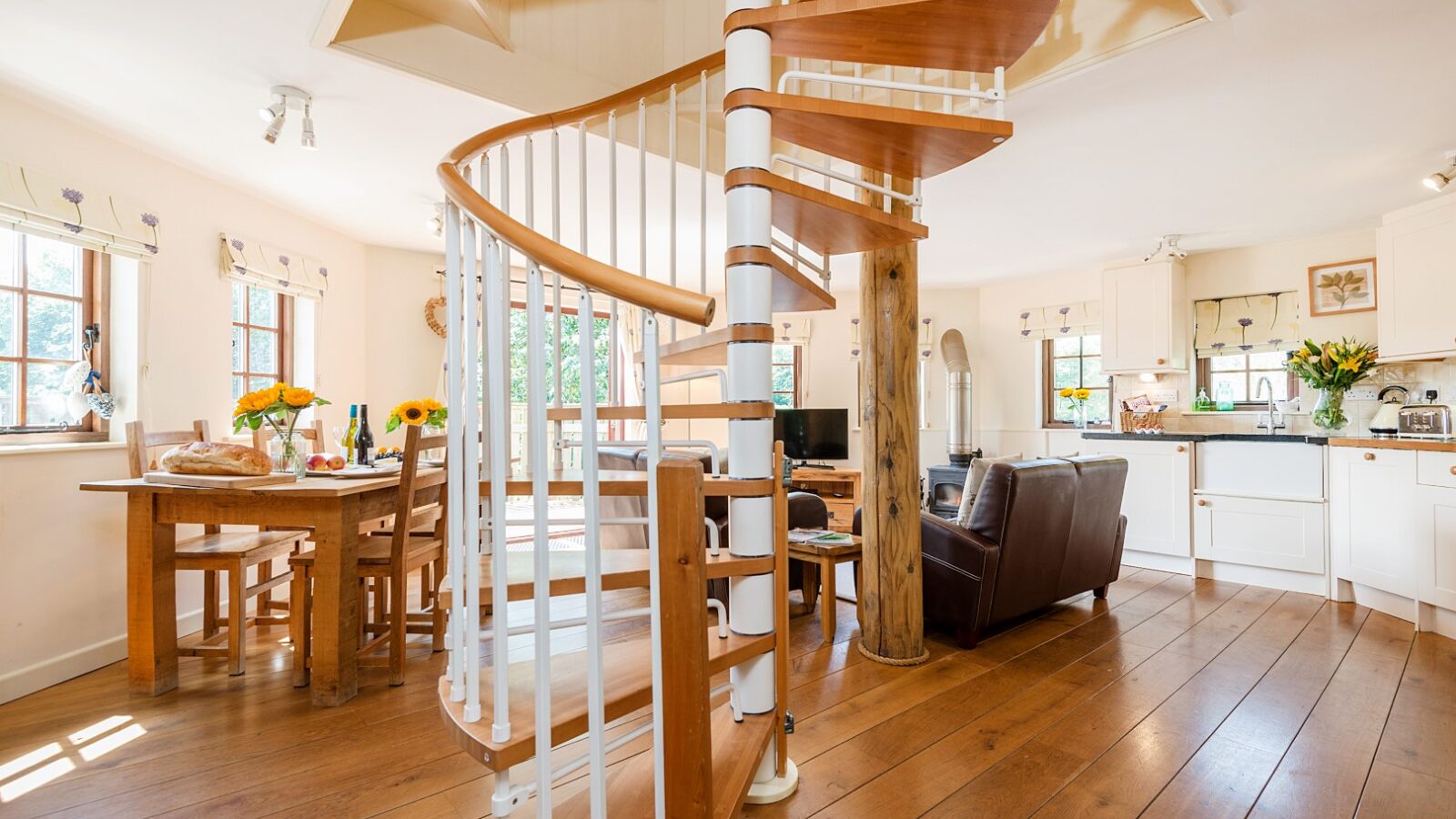 A spiral wooden staircase in a cozy Lavender Hill open-plan living area with a dining table, chairs, and kitchen in the background evokes the charm of luxurious treehouses.