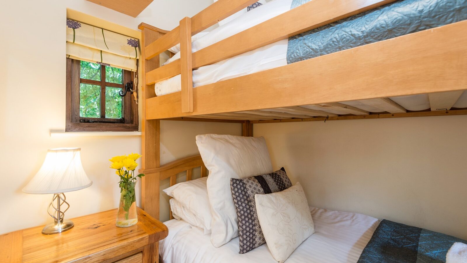 Cozy bedroom reminiscent of Lavender Hill's charming treehouses, featuring a wooden bunk bed, decorative pillows, side table, lamp, and flowers by a small window.